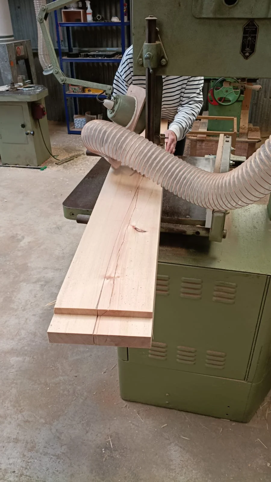 A person operating a woodworking bandsaw machine cutting a long wooden plank, with wood shavings on the floor in a workshop.