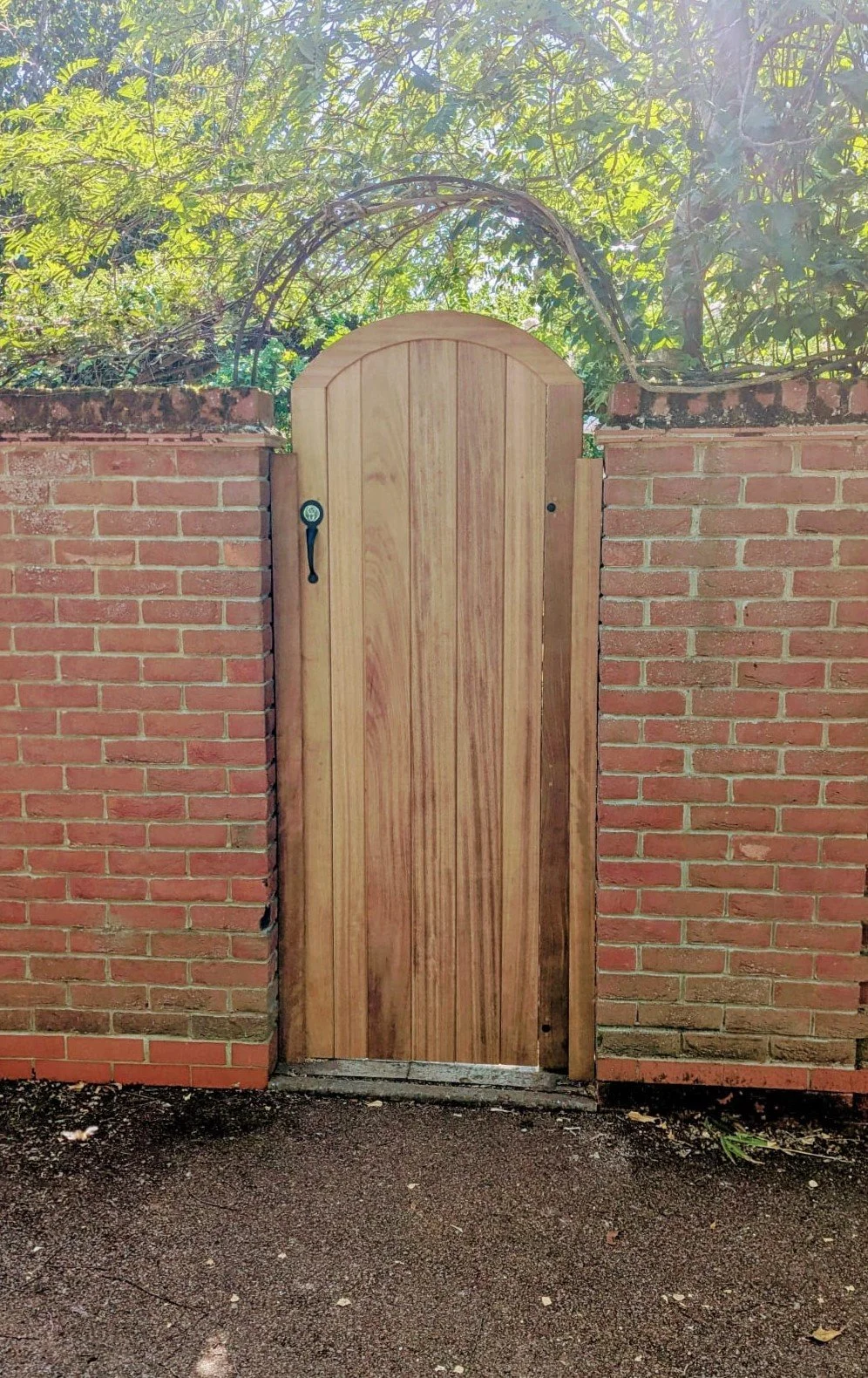 A wooden gate with an arched top set into a brick wall. The gate has a black handle and a circular hook, with greenery and trees visible behind the wall.