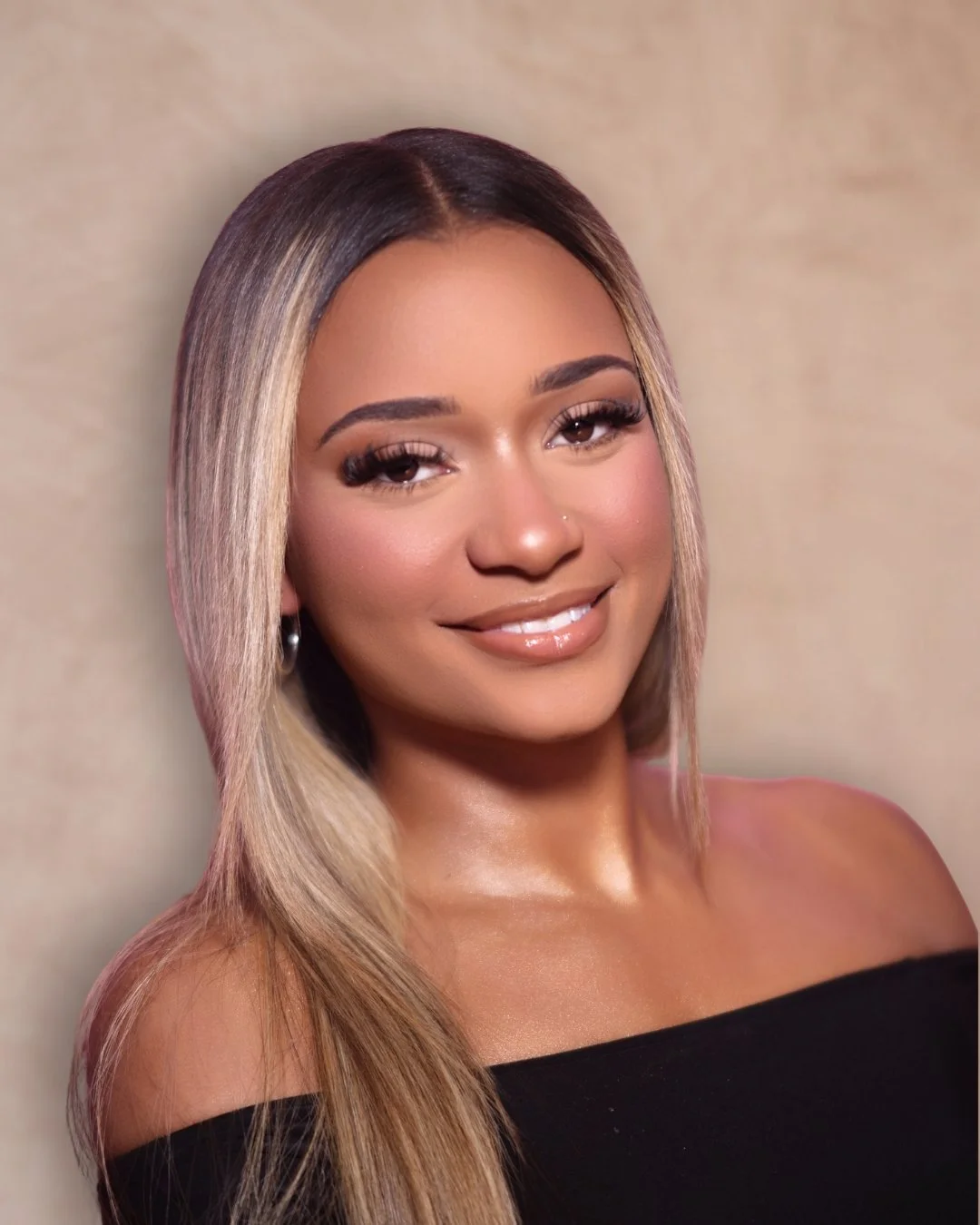 A smiling woman with long, blonde hair, wearing a black off-the-shoulder top, posing against a neutral background.