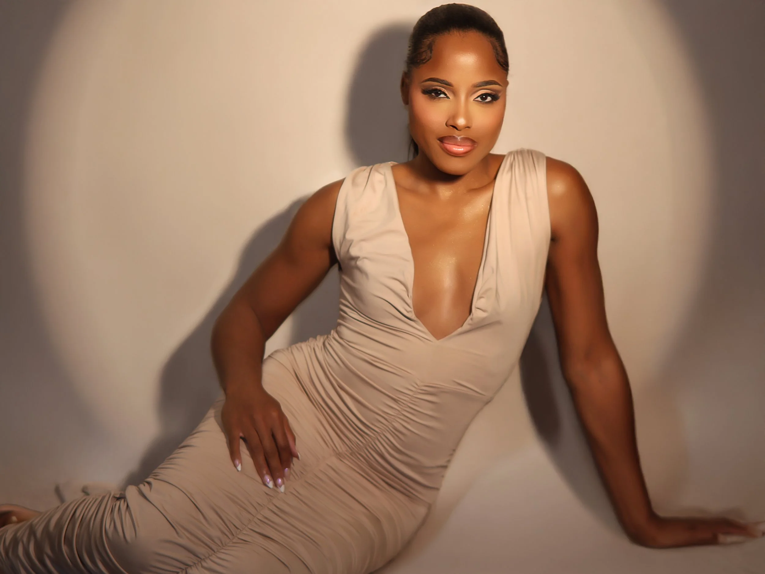 A woman with styled braids decorated with white beads, wearing makeup with glossy lips, and a gold necklace, dressed in a white sleeveless top with black accents, standing against a plain brown background.