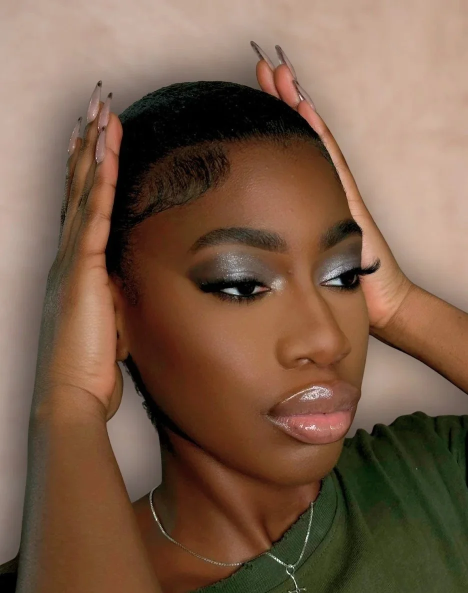 A portrait of a woman with short, styled hair, shimmering eyeshadow, and glossy lips, wearing a green top, rings, and a necklace with a cross pendant.