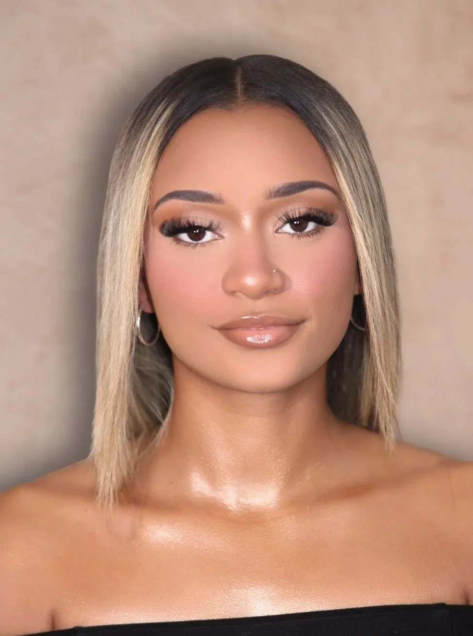 Close-up of a smiling woman with reddish-brown braided hair, makeup, and tattoos, wearing a black top, gold earrings, and layered necklaces, standing against a neutral background.