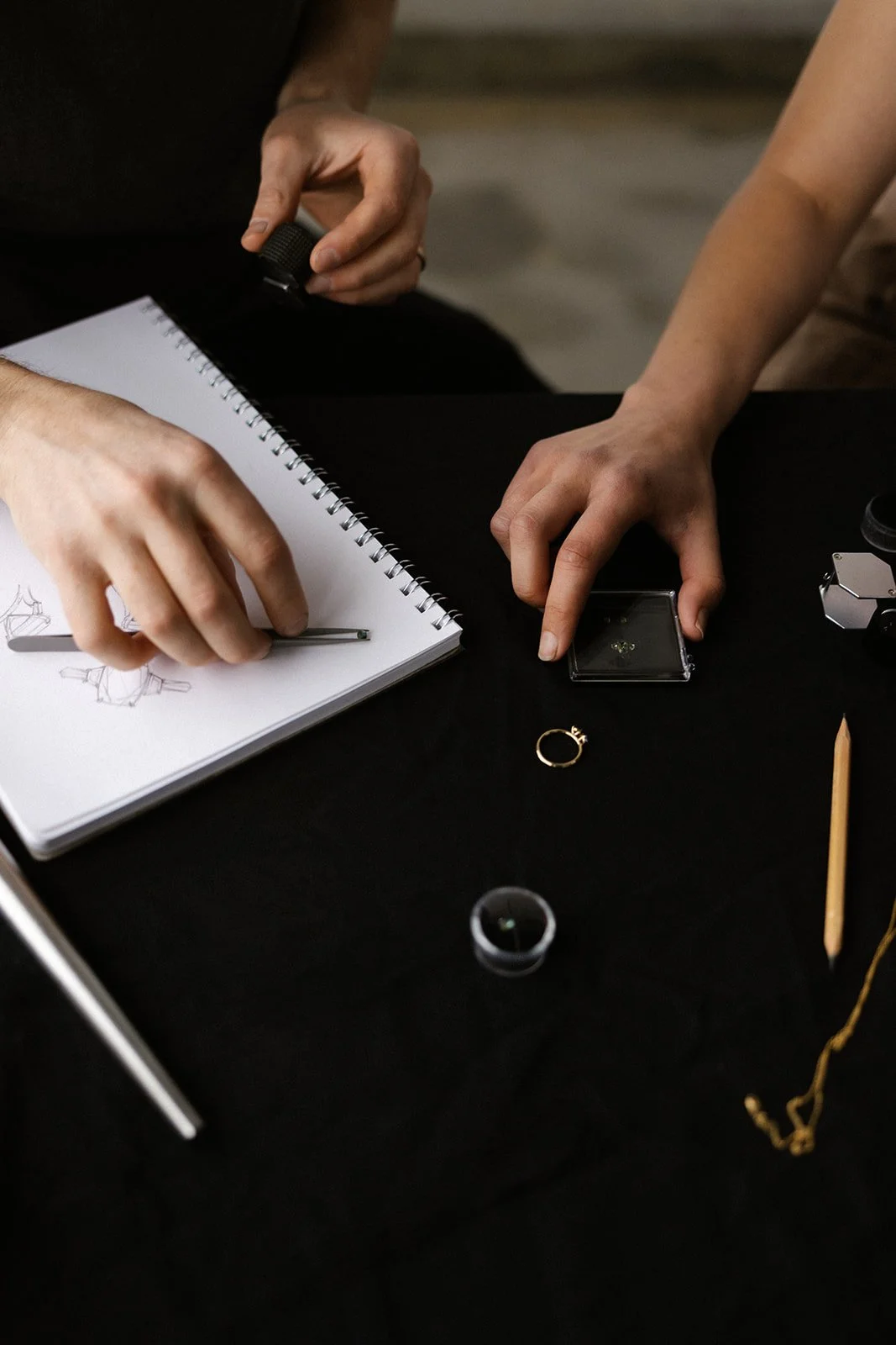 A man with dark hair looking through a magnifying glass at a jewelry piece on a table with sketchbook, markers, and boxes in front of him, against a background of a light-colored brick wall.