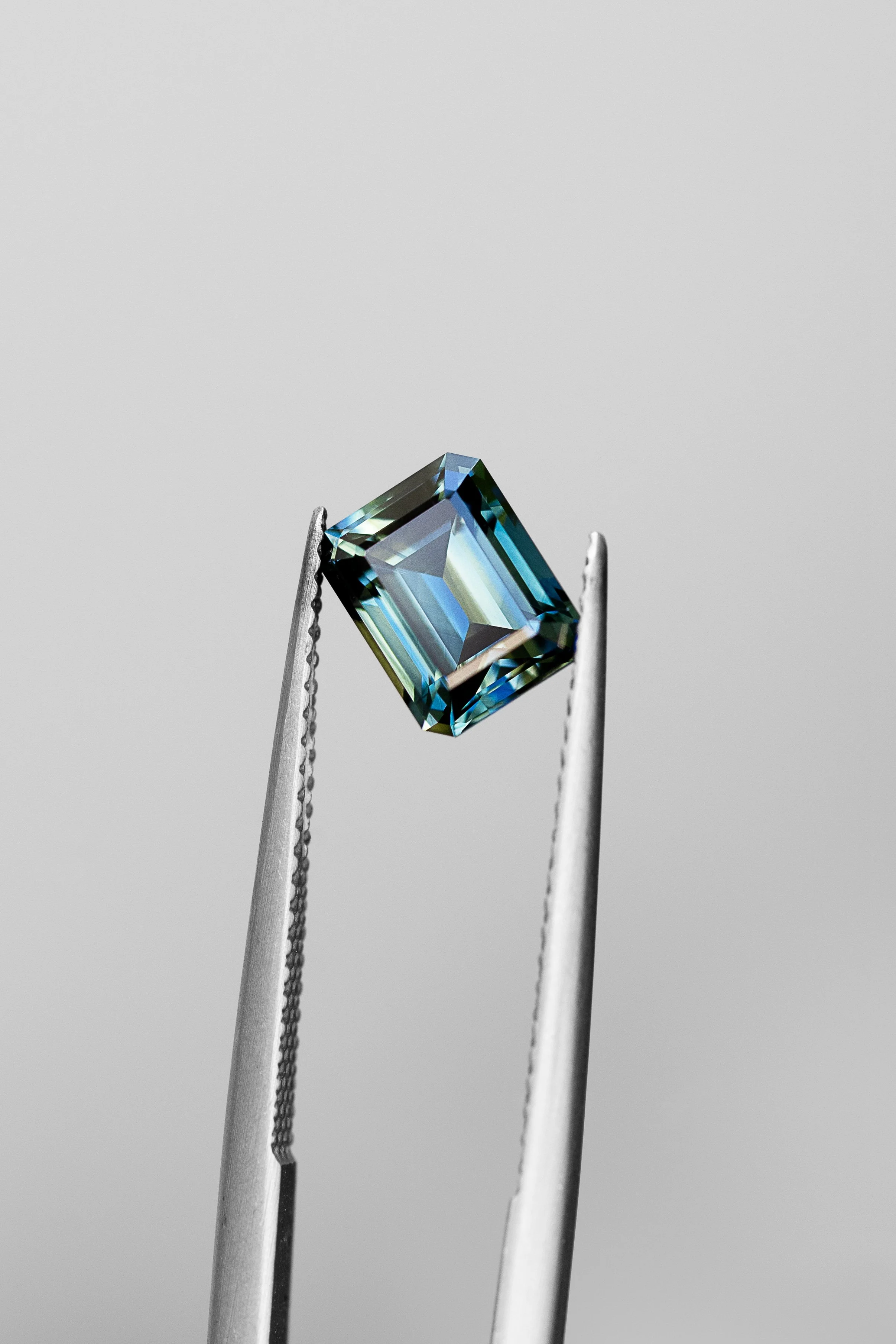 A pair of tweezers holding a rectangular blue gemstone against a plain background.