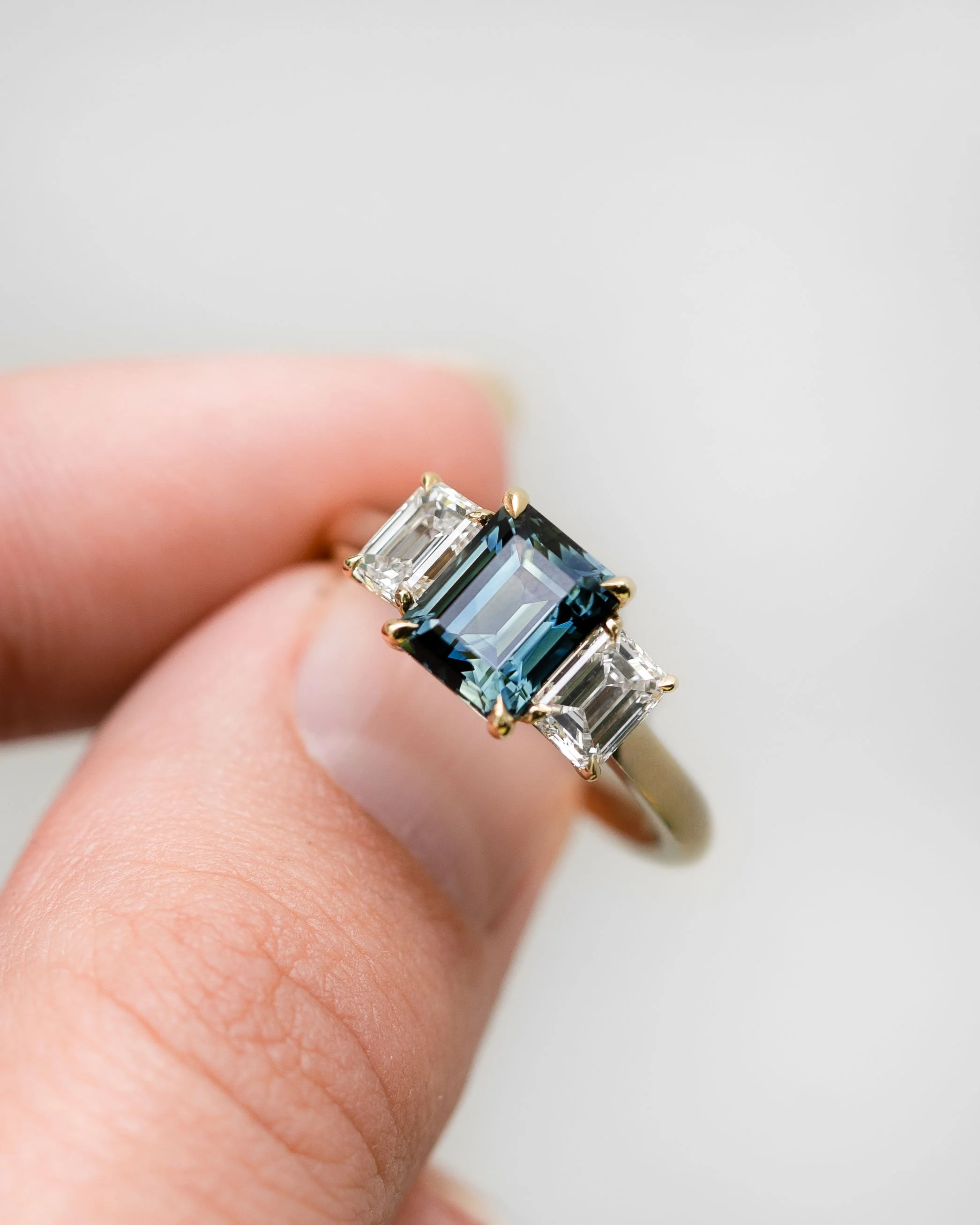 Close-up of a person's hand with a gold ring featuring a large blue sri lankan sapphire and small diamonds on a band. The person has a small tattoo of a square with a line inside on the finger. The hand rests on light-colored fabric.
