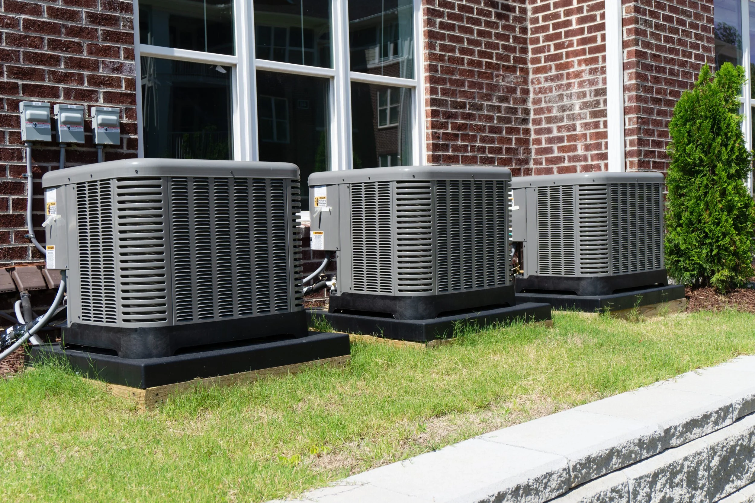 Three outdoor air conditioning units installed on a lawn outside a brick house with a window and a small bush.