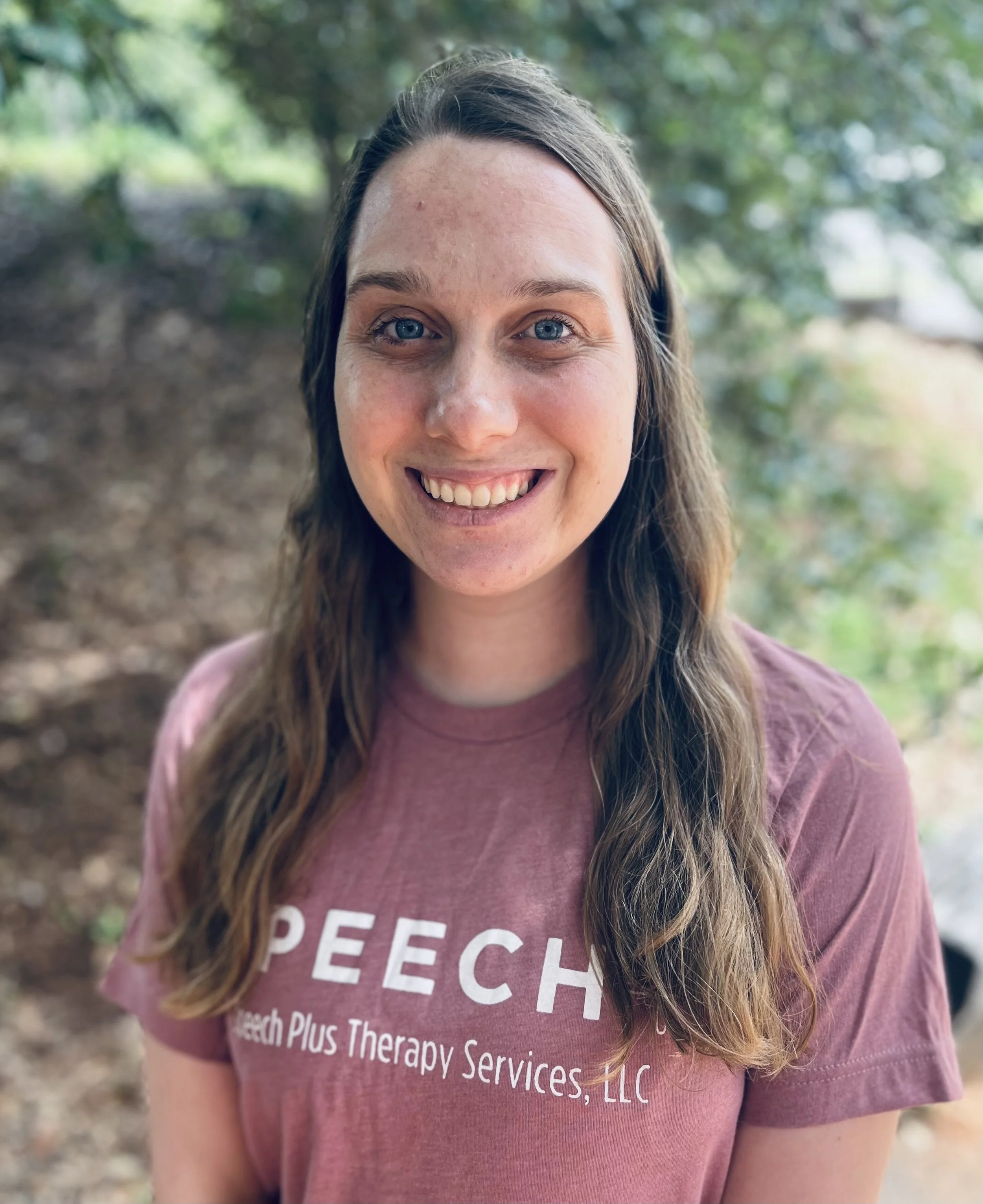 A young woman with long wavy brown hair and blue eyes smiling outdoors in a natural setting, wearing a maroon T-shirt with the text 'SPEECH' and 'Speech Plus Therapy Services, LLC'.
