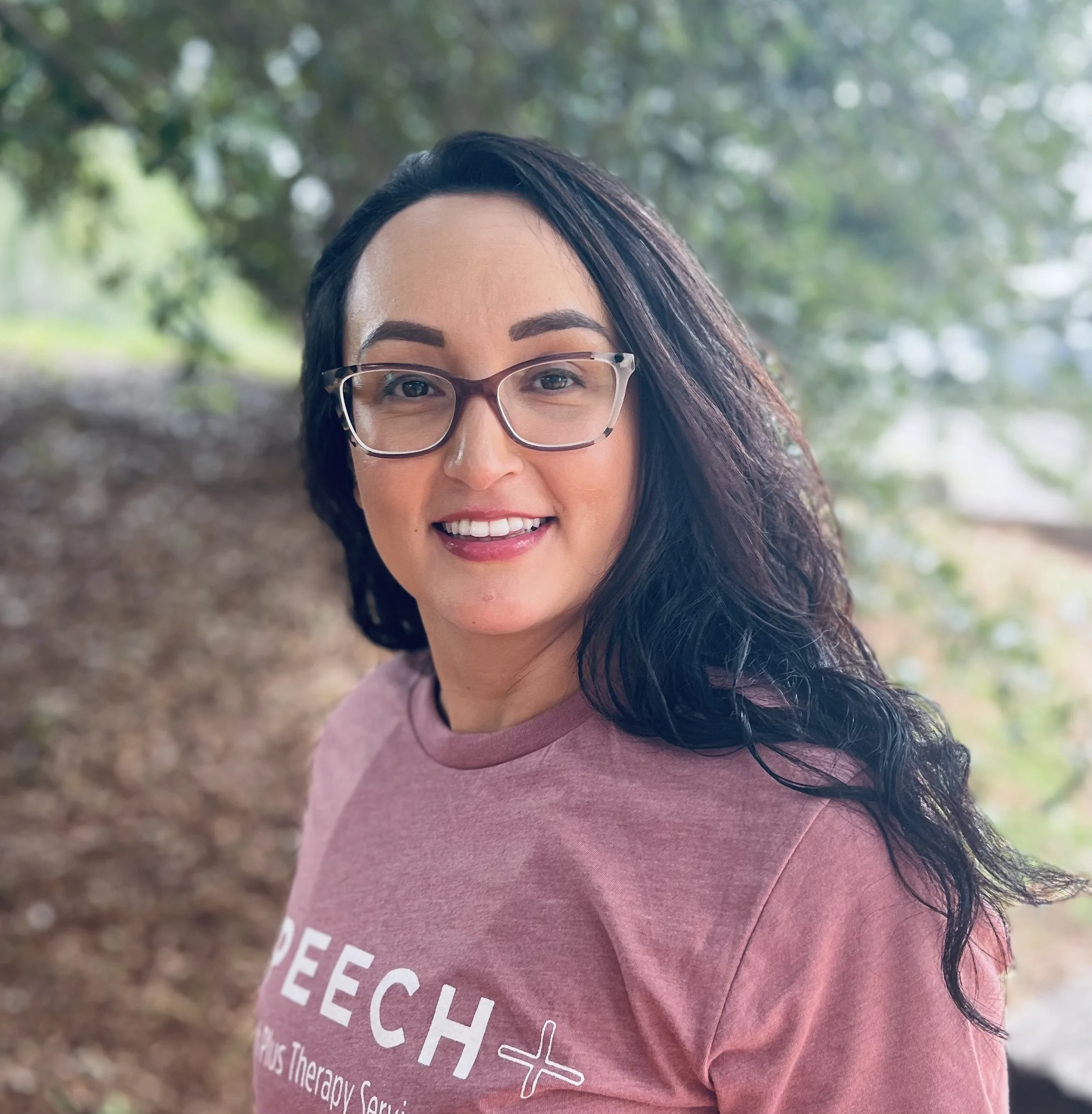 A woman with black hair, glasses, and a pink t-shirt standing outdoors with trees and foliage in the background.