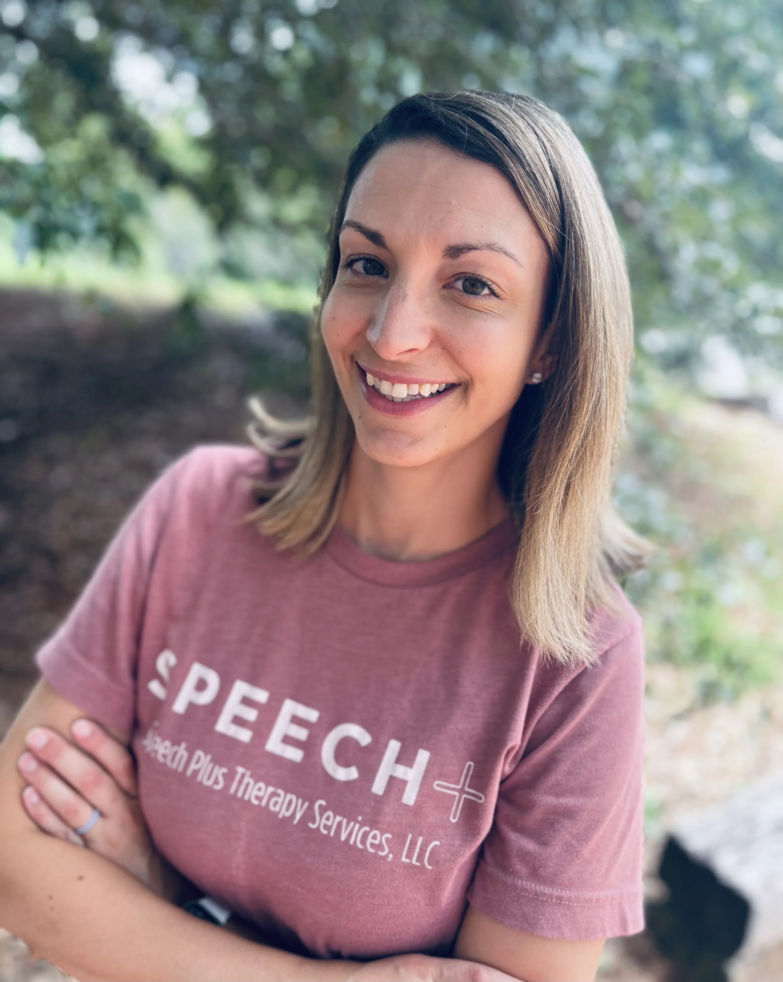 A smiling young woman with shoulder-length light brown hair, wearing a pink t-shirt with white text that reads 'SPEECH Speech Plus Therapy Services, LLC,' standing outdoors with trees and greenery in the background.
