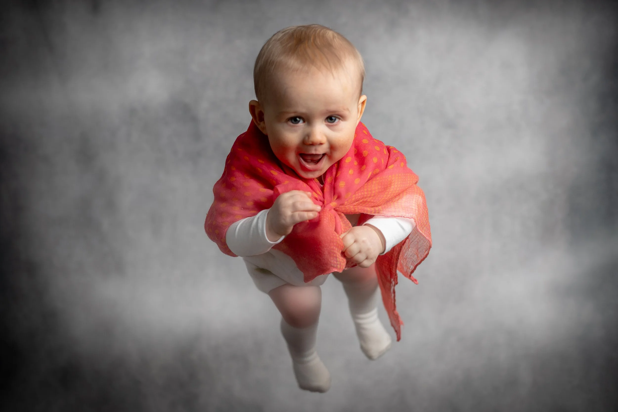 Iloinen vauva, joka on pukeutunut valkoisiin vaatteisiin ja punavihreään huiviin, lentää harmaalla taustalla ja katsoo kameraan.