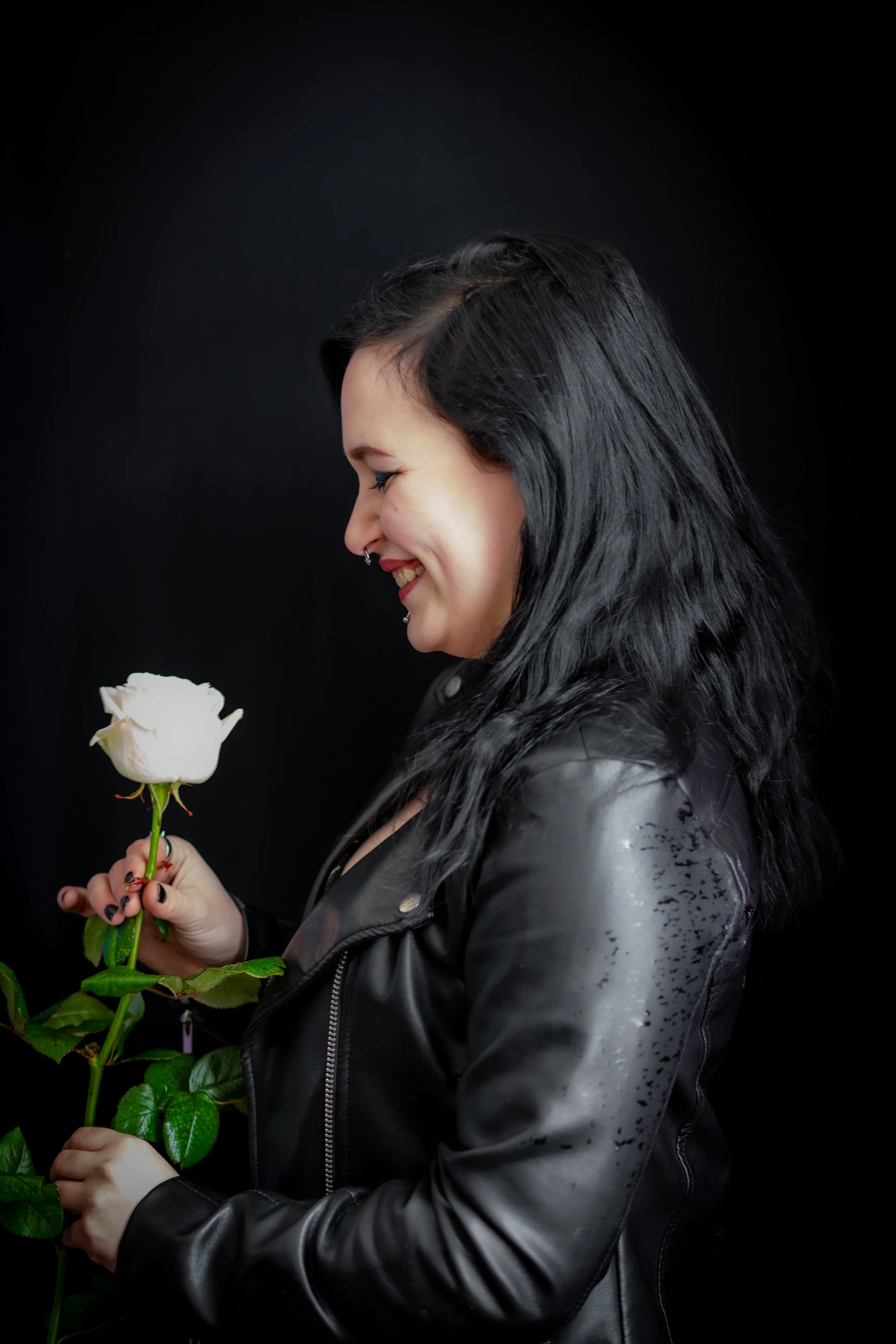 Nainen hymyilee ja katsoo valkoista ruusua, jonka hän pitää kädessään, taustalla musta.
