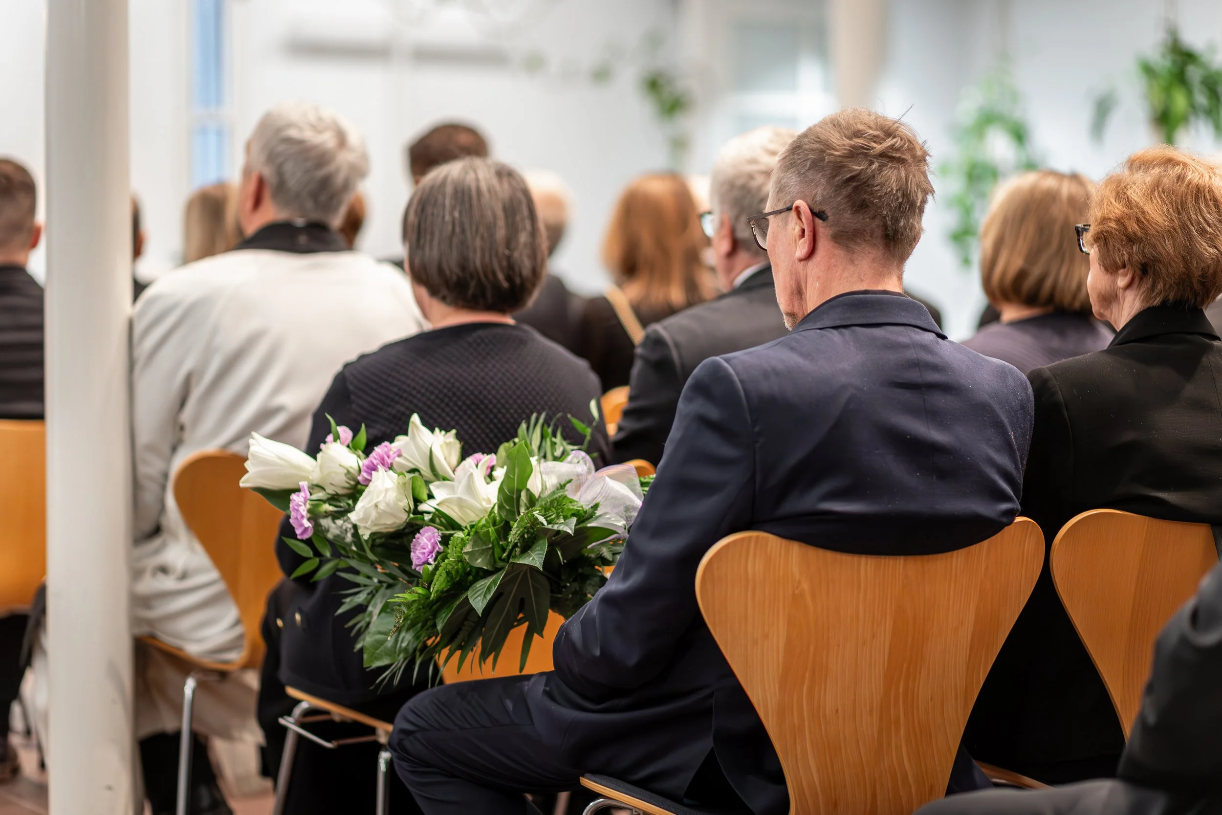 Ihmiset istuvat istuimilla seminaarissa tai kokouksessa, ja sisältä näkyy kukkakimppu pöydällä.