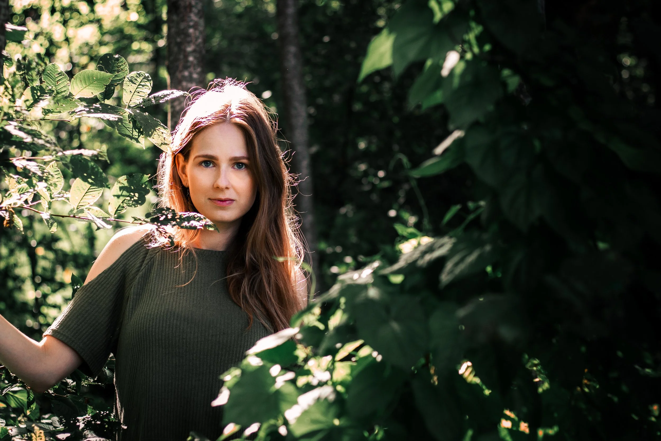 Nuori nainen vihreästä puutarhasta tai metsästä, katsoo kameraan, takana runsaasti vihreitä lehtiä ja puita, kesäinen päivä.