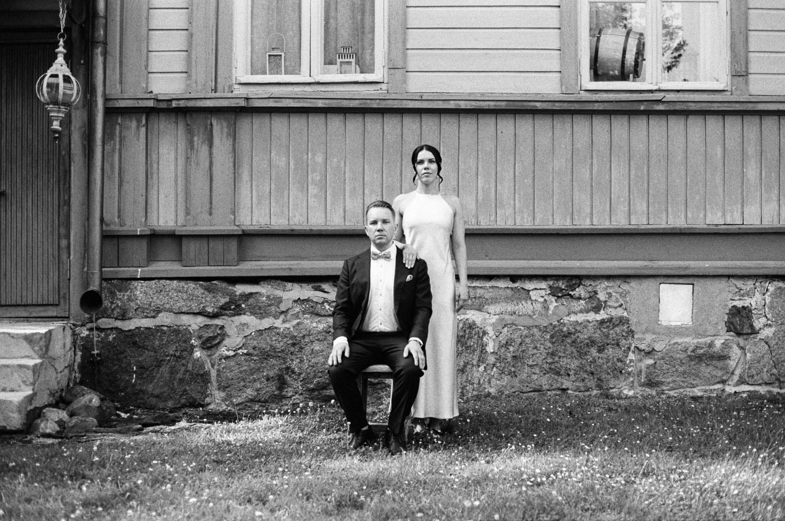 Mustavalkoinen valokuva pariskunnasta, mies istuu tuolilla ja nainen seisoo hänen takanaan, rakennuksen ulkoseinä taustalla.