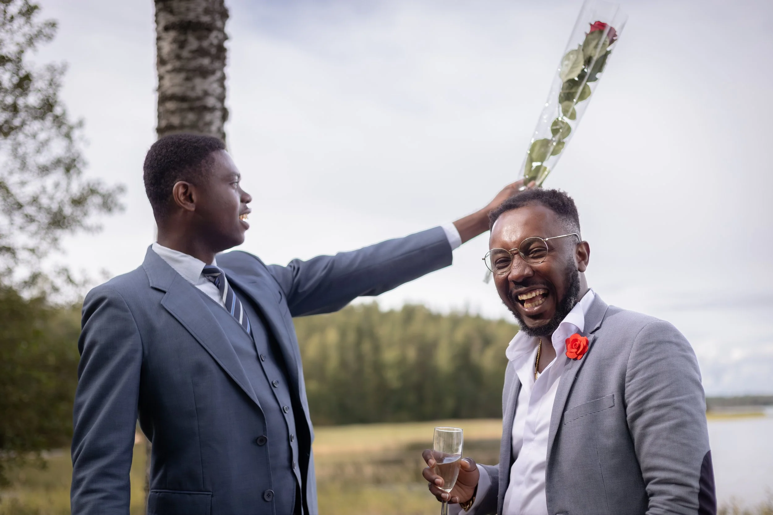 Kaksi miestä juhlivassa ulkona, toinen nauraa ja toinen ojentaa ruusun päähän, toinen pitää lasillista juomaa.