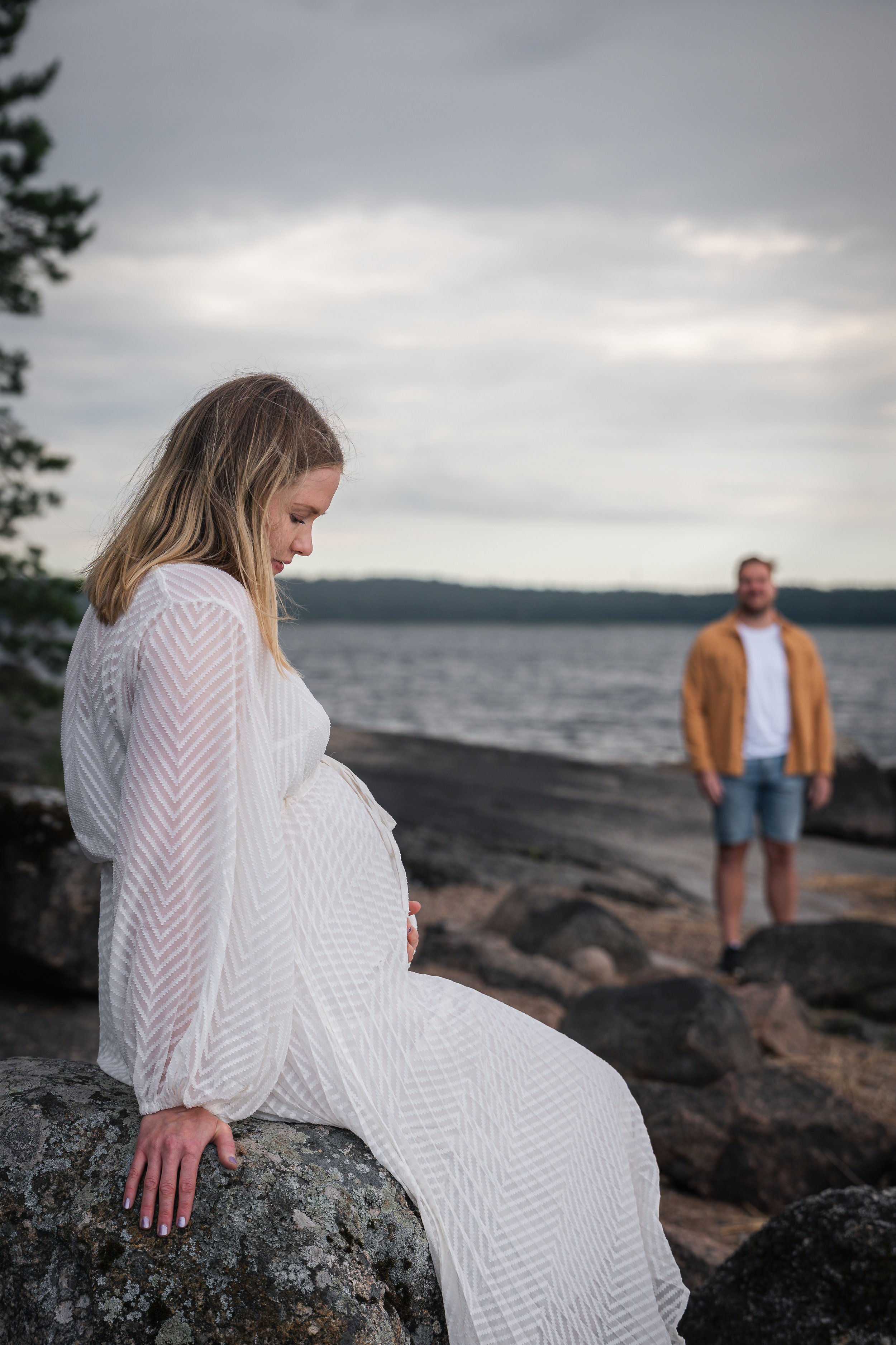 Raskaana oleva nainen istuu kalliolla rannalla, taustalla näkyy mies. Tummat pilvet ja vesistö näkyvät taustalla.