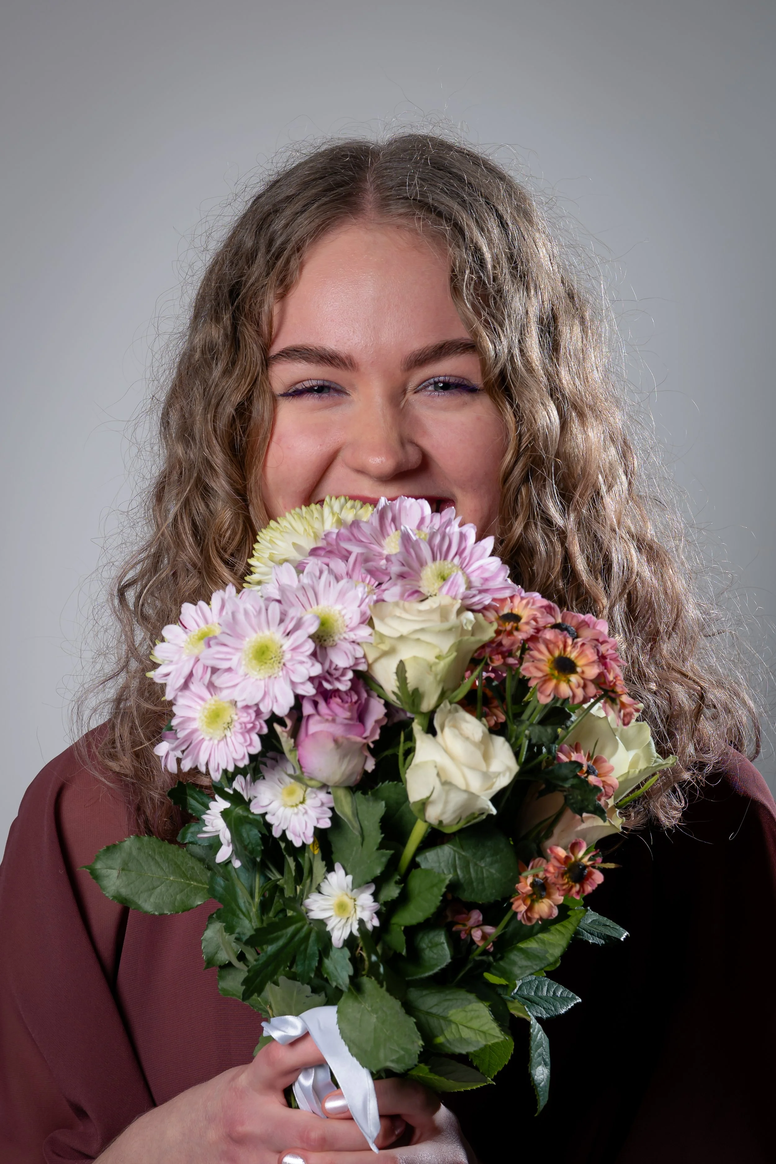 Nainen hymyilee ja pitää kimppua erilaisia kukkia, kuten pionteja, kehäkukkia, valkoisessa sidottuna nauhalla.