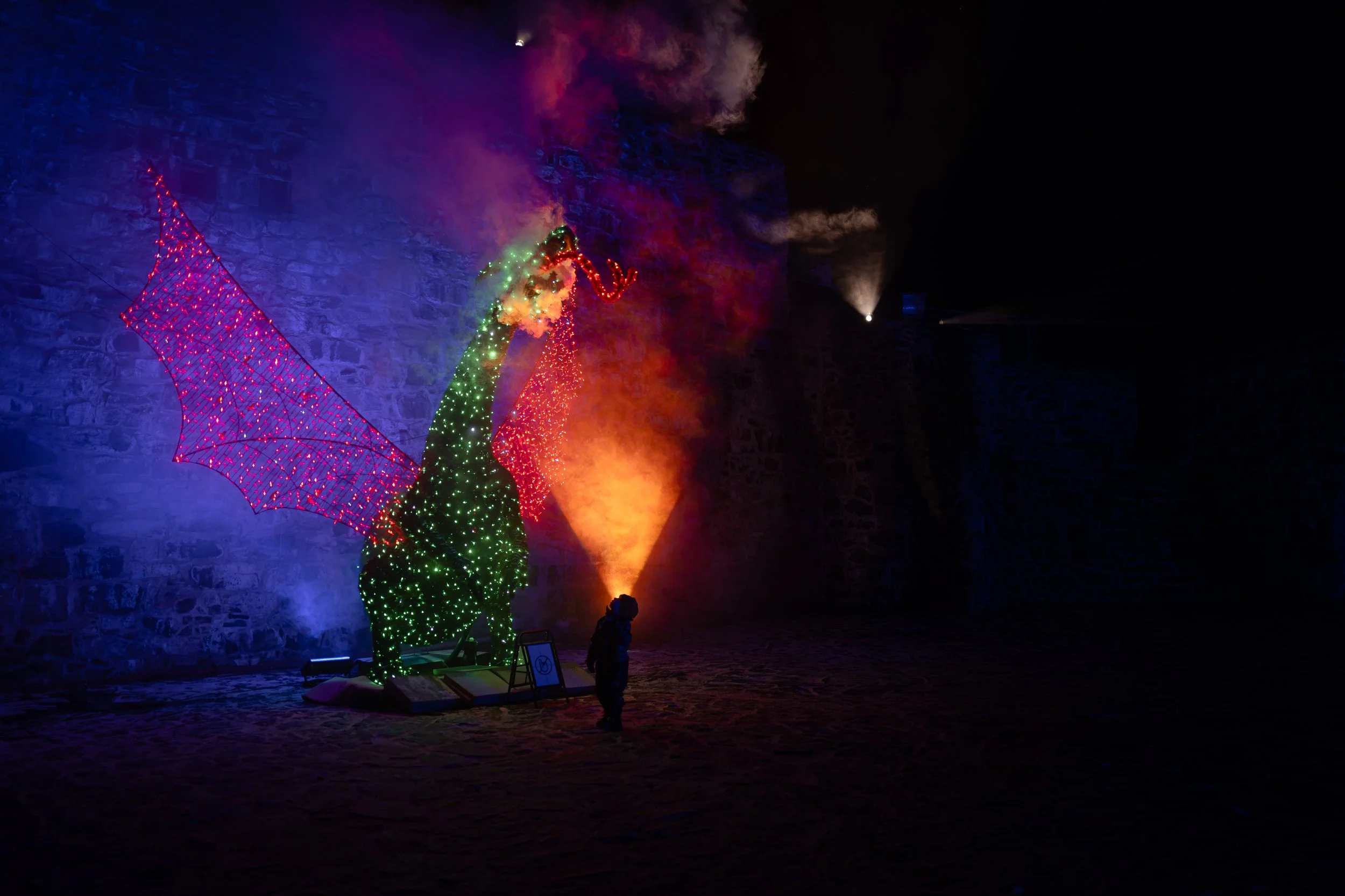 Vihreä lohikäärme, jossa on punaiset siivet ja risteilevät valot, roikkuu kivi- ja betoniarkkitehtuurin edessä yöllä, taustalla palaa sinistä ja punaista valoa merkin ja savun muodossa.