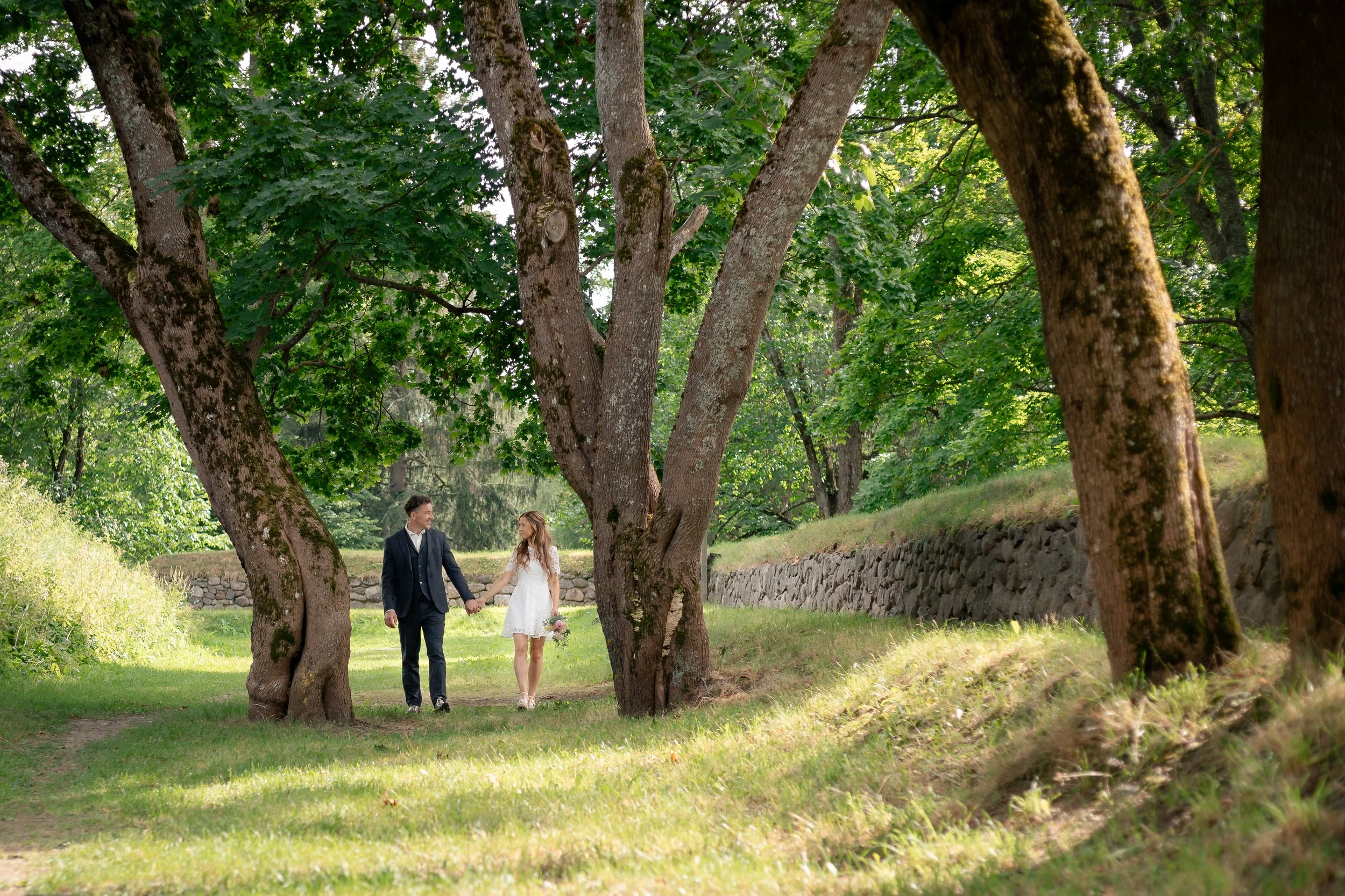 Pariskunta kävelee käsi kädessä puistossa, ympärillä vihreitä puita ja niittyä.