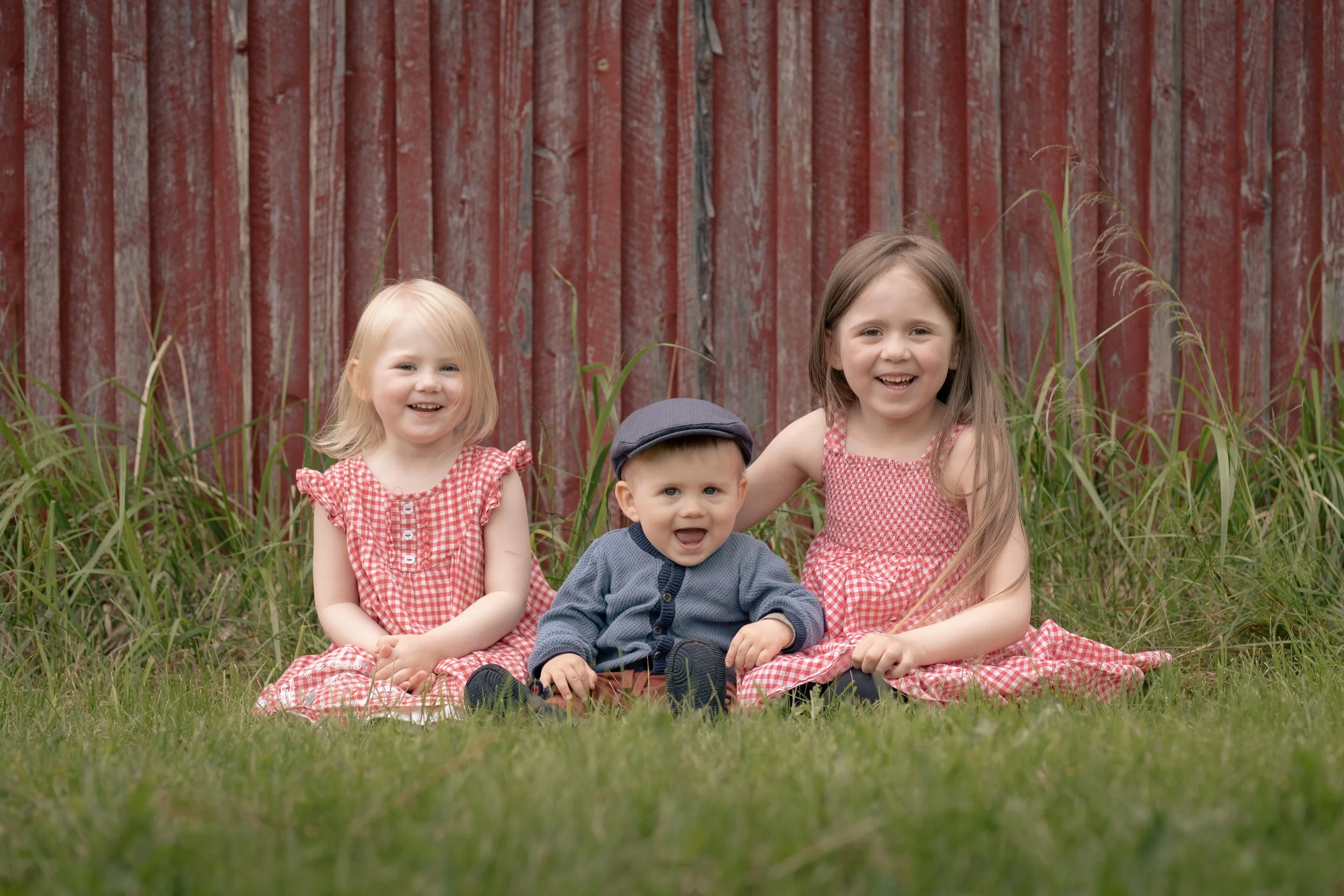 Kolme lasta istuu nurtsilla pihan nurmikolla punaisen aidan edessä.