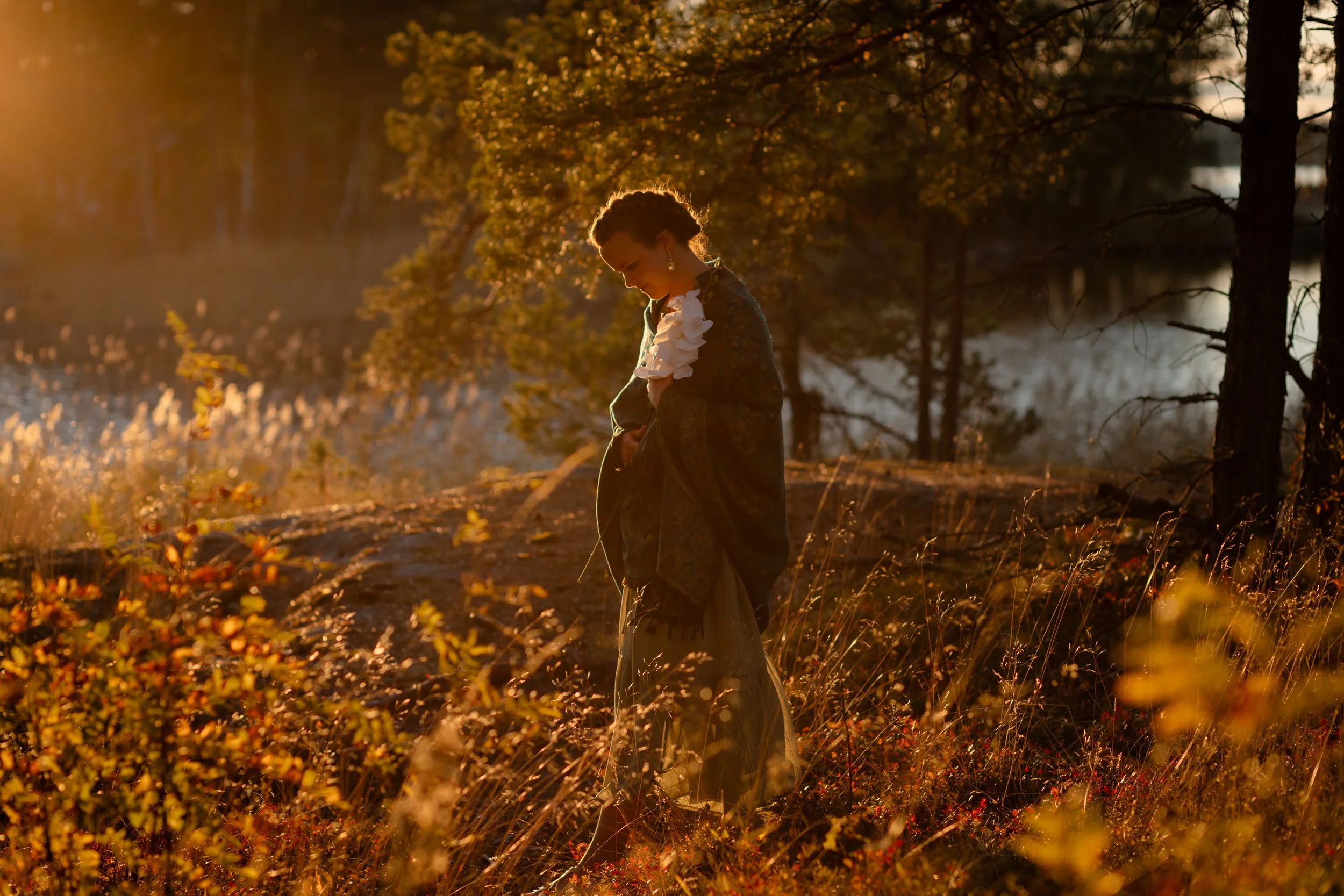Nainen pukeutunut vanhanaikaiseen vaatteeseen seisoo luonnon keskellä auringon laskiessa, läheisellä järvellä ja ympäröivissä metsissä.