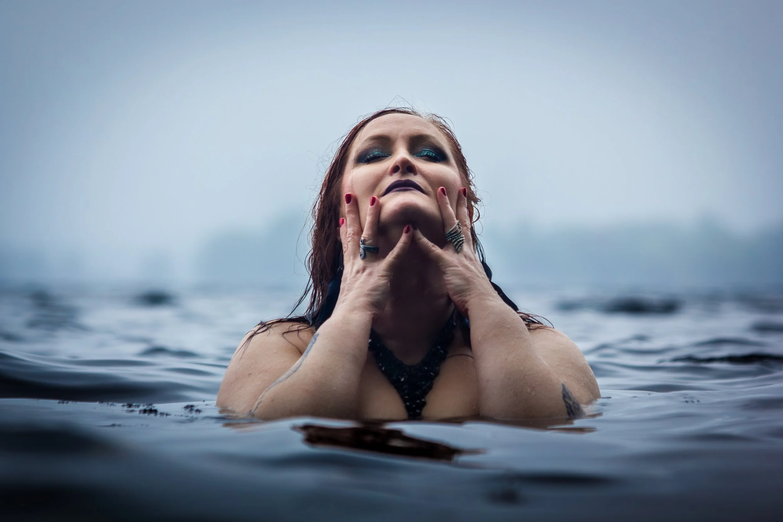Vesistöön upotettu nainen, jonka kädet tukevasti hänen kasvoillaan, ja taustalla harmaa taivaskuva.