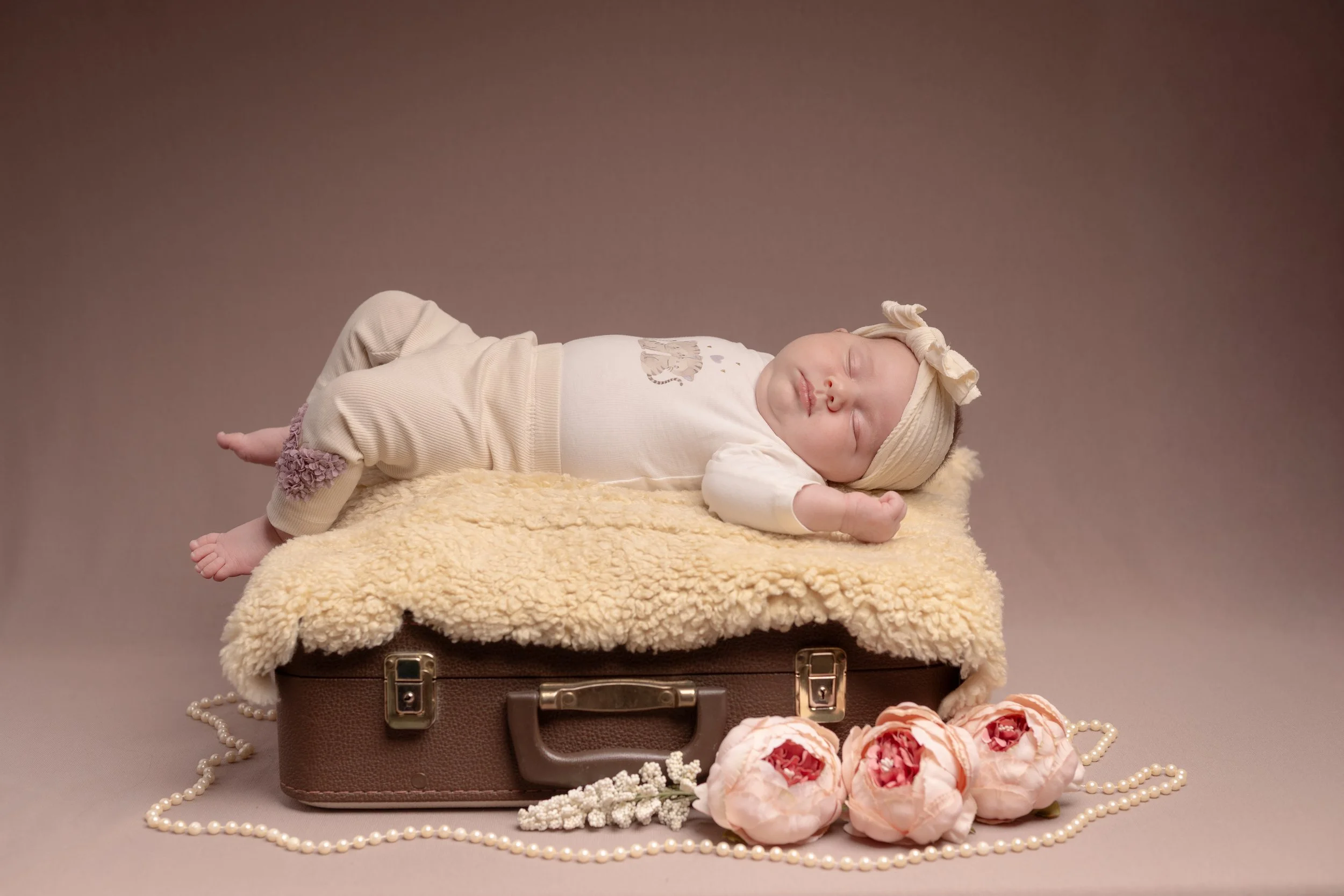 Vauva makaa suloisella alustalla, jossa on kellastunut villahuopa, saaden kuvaa kuin lelukuvasta. Iholla on valkoinen paita ja beigehihat, ja hänellä on päässään keltainen rusetti.