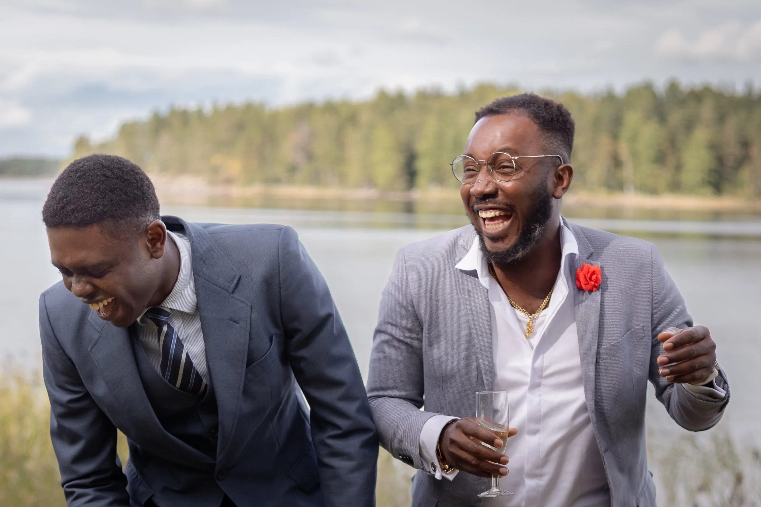 Kaksi miestä juhlissaan puvut päällä, nauttien juomia ulkona järven rannalla, nauraen ja hymyillen.