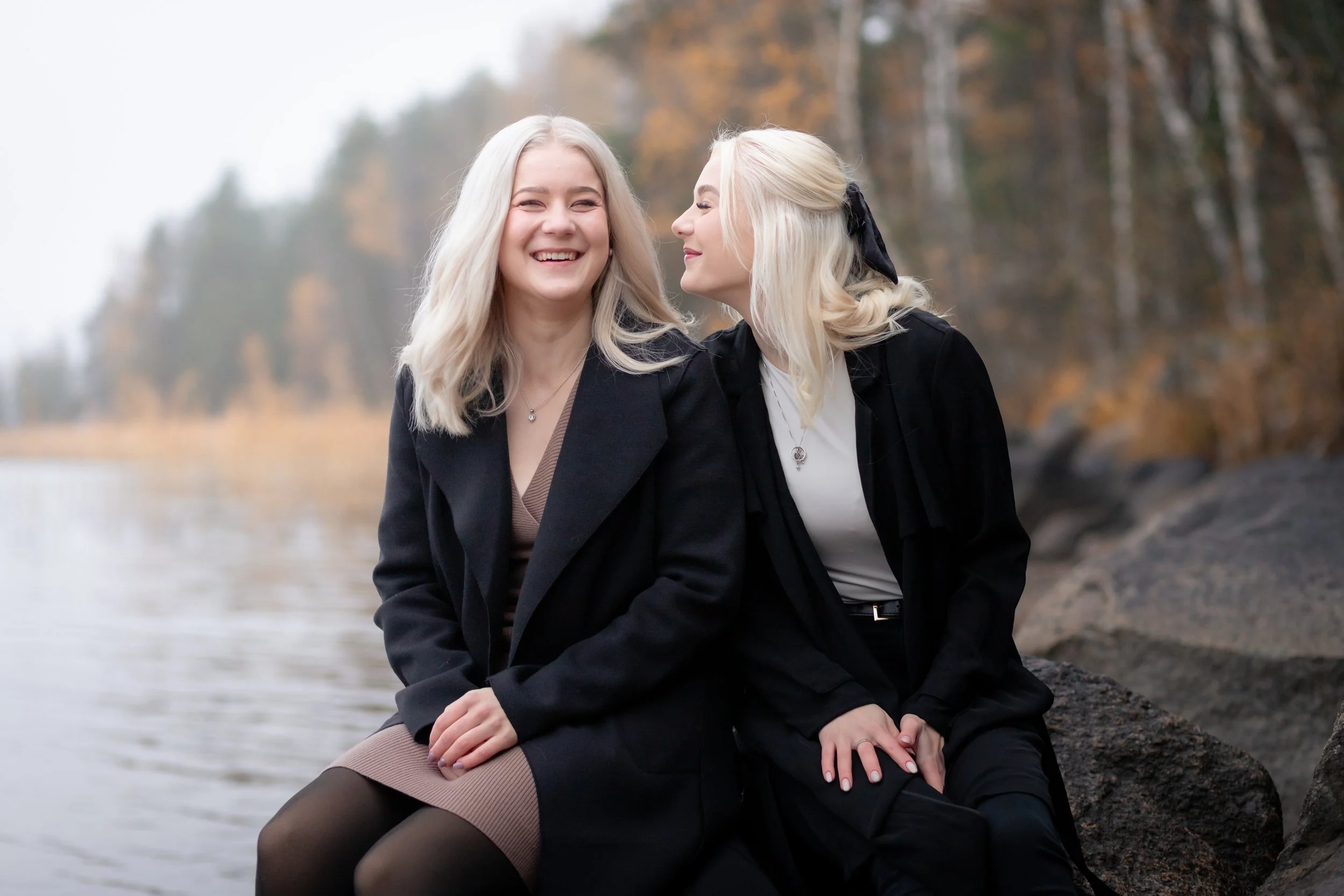 Kaksi naista istuu rantakivillä ja nauraa, taustalla syksyinen metsä ja järvi.
