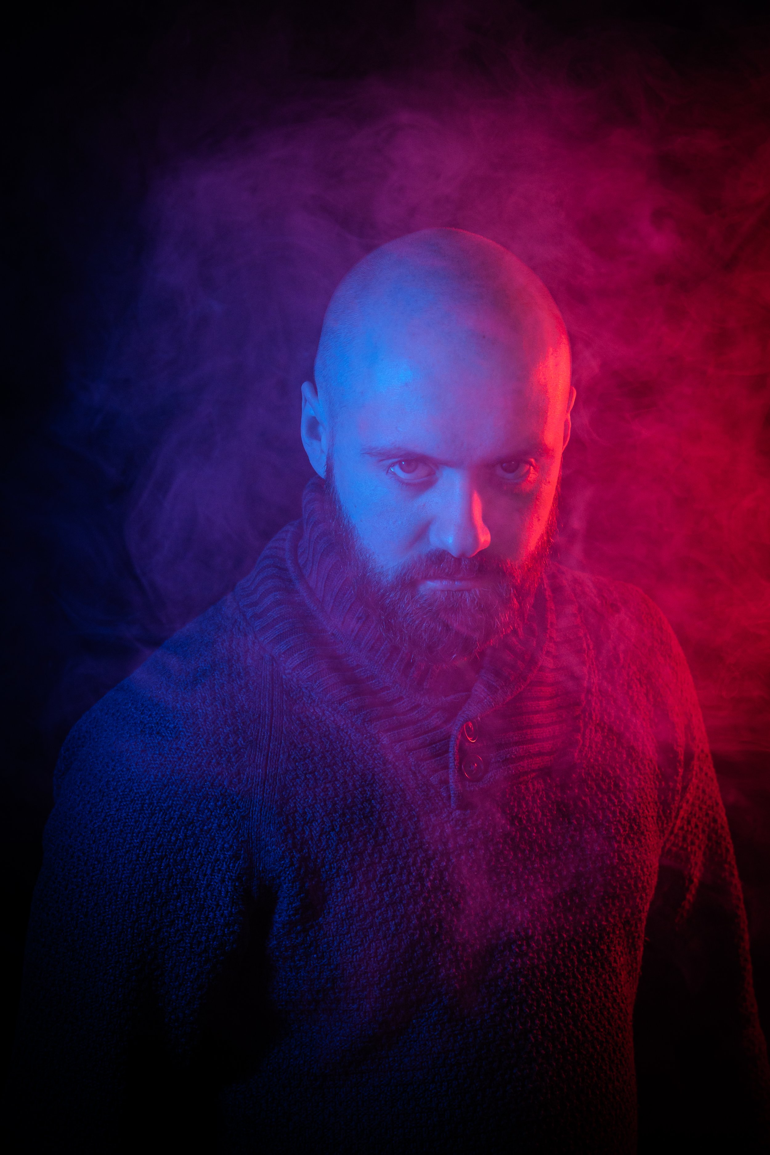 Tumma kuva jossa mies katsoo tuimasti kohti kameaa, taustalla sinistä ja punaista valoa sekä savua.