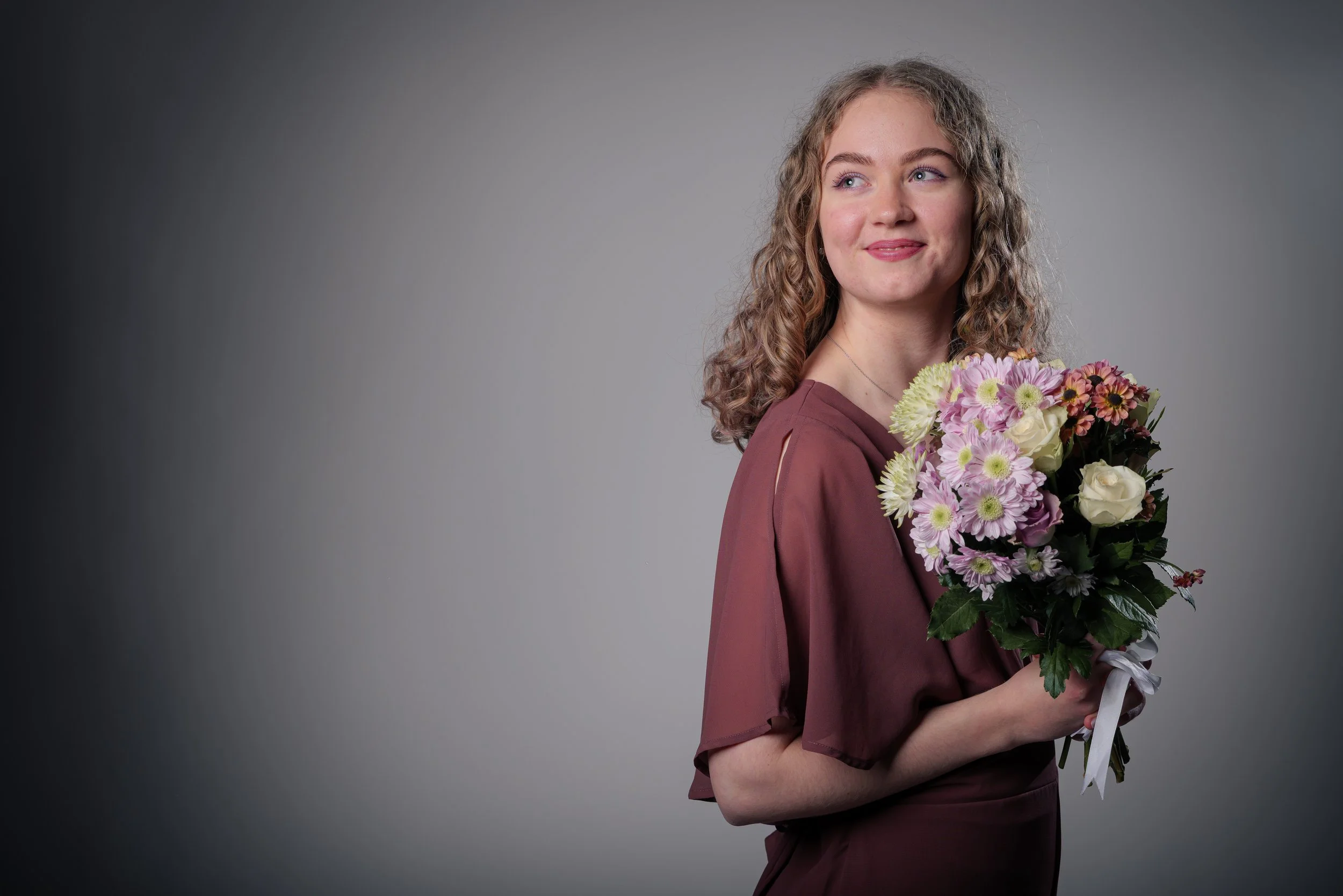 Nuori nainen, jossa kiharat hiukset ja mekko, pitää kukkakimppua, taustalla harmaa.