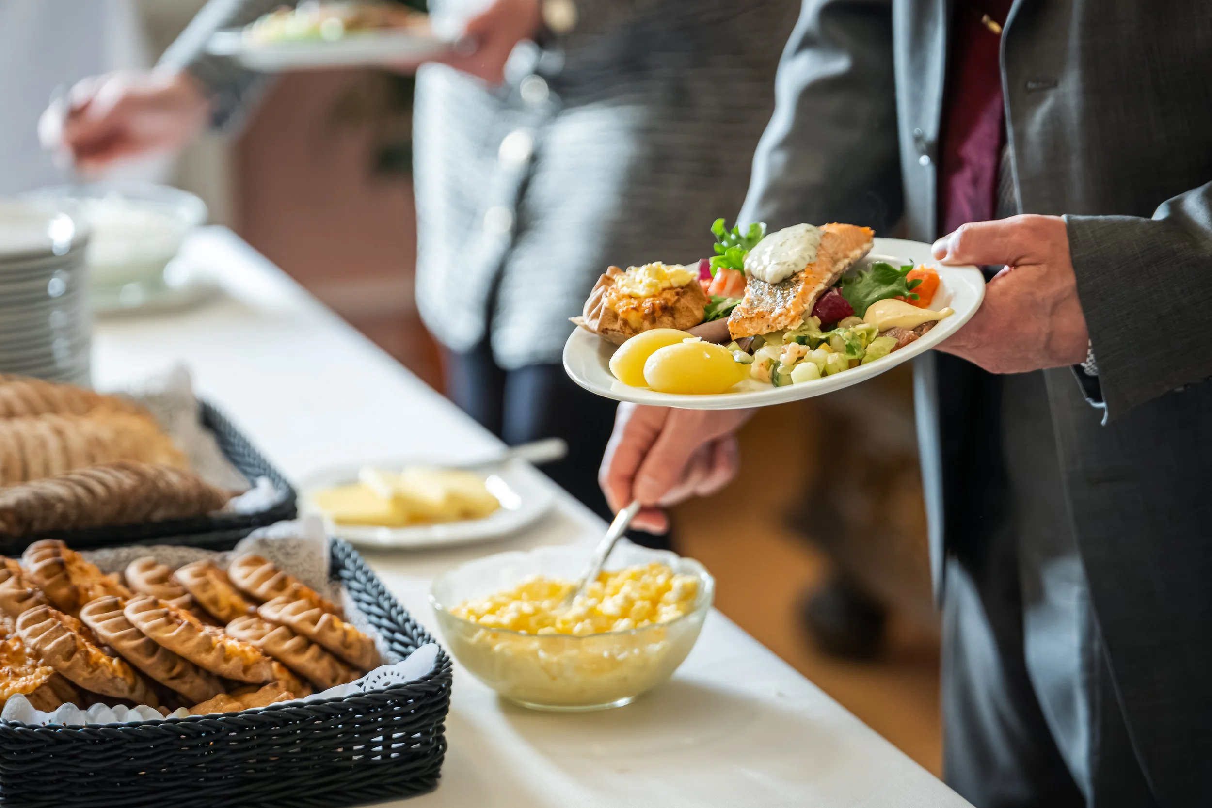 Henkilö tarjoilee lautaselta erilaisia salaatteja ja kaloja buffet-pöydässä, taustalla on kasa keksejä ja perunasalaattia.