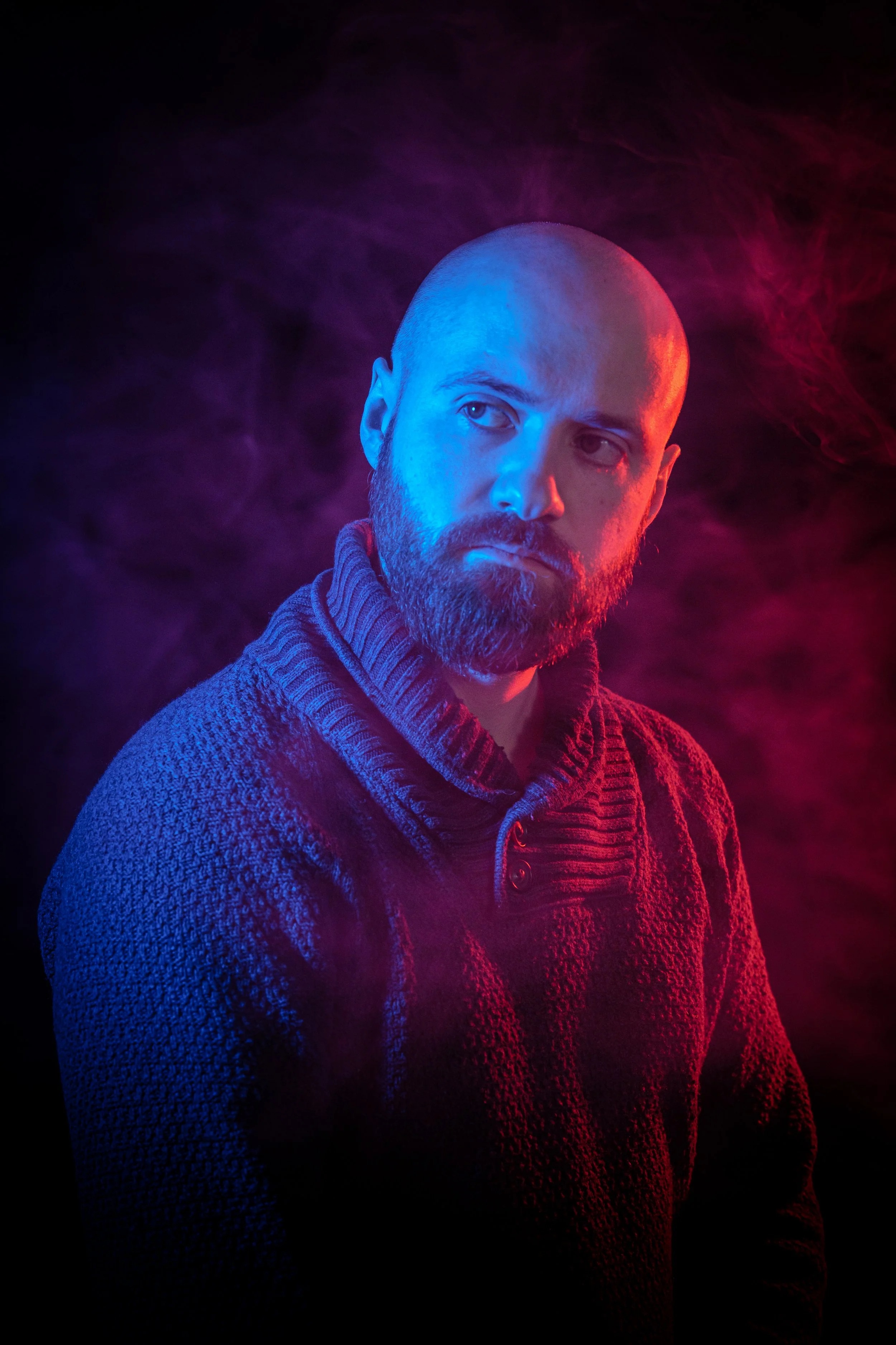 Mies katselee kameraa vasten, tausta ja savu neon-valaistuksella, pusero ja parta, värivaloilla