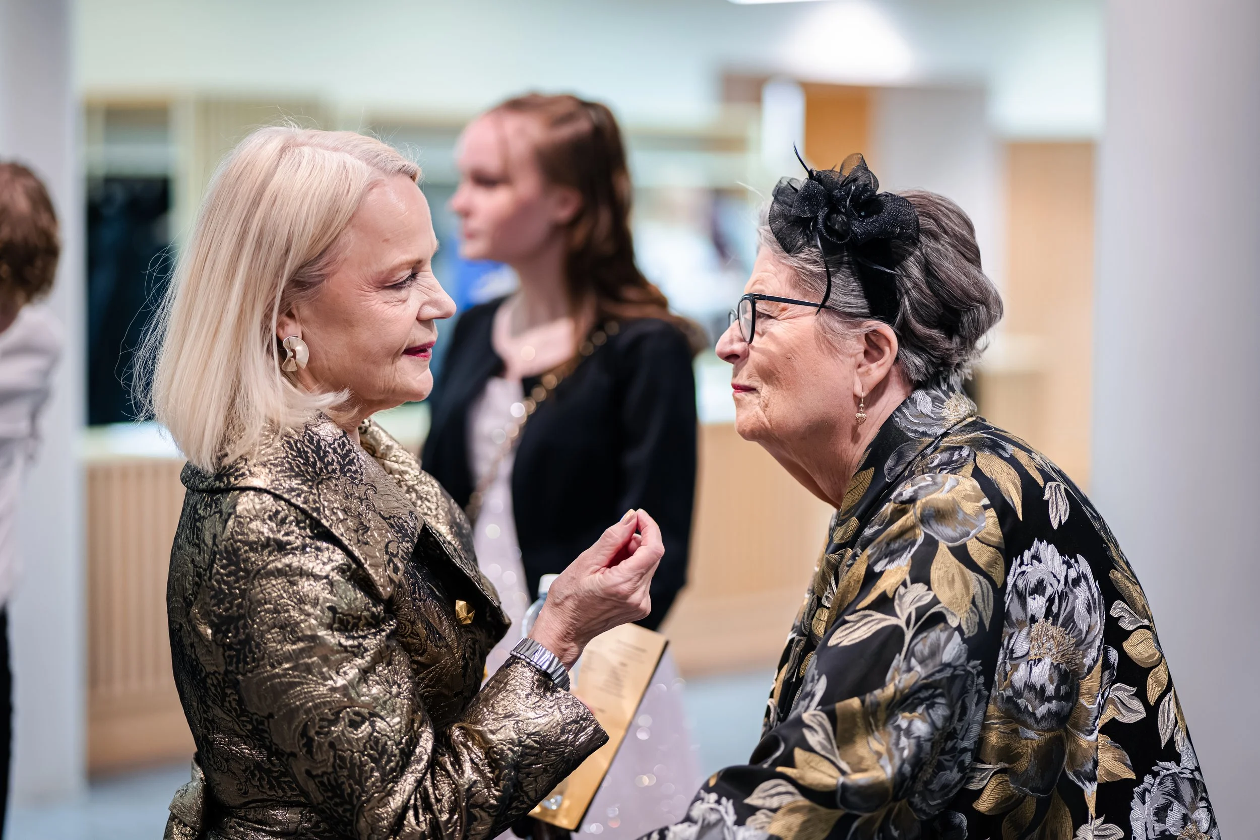 Kaksi hyvin pukeutunutta naista keskustelee lähekkäin tapahtumassa, toinen nainen pitää toisen kättä ja heidän ilmeensä ovat ystävälliset.