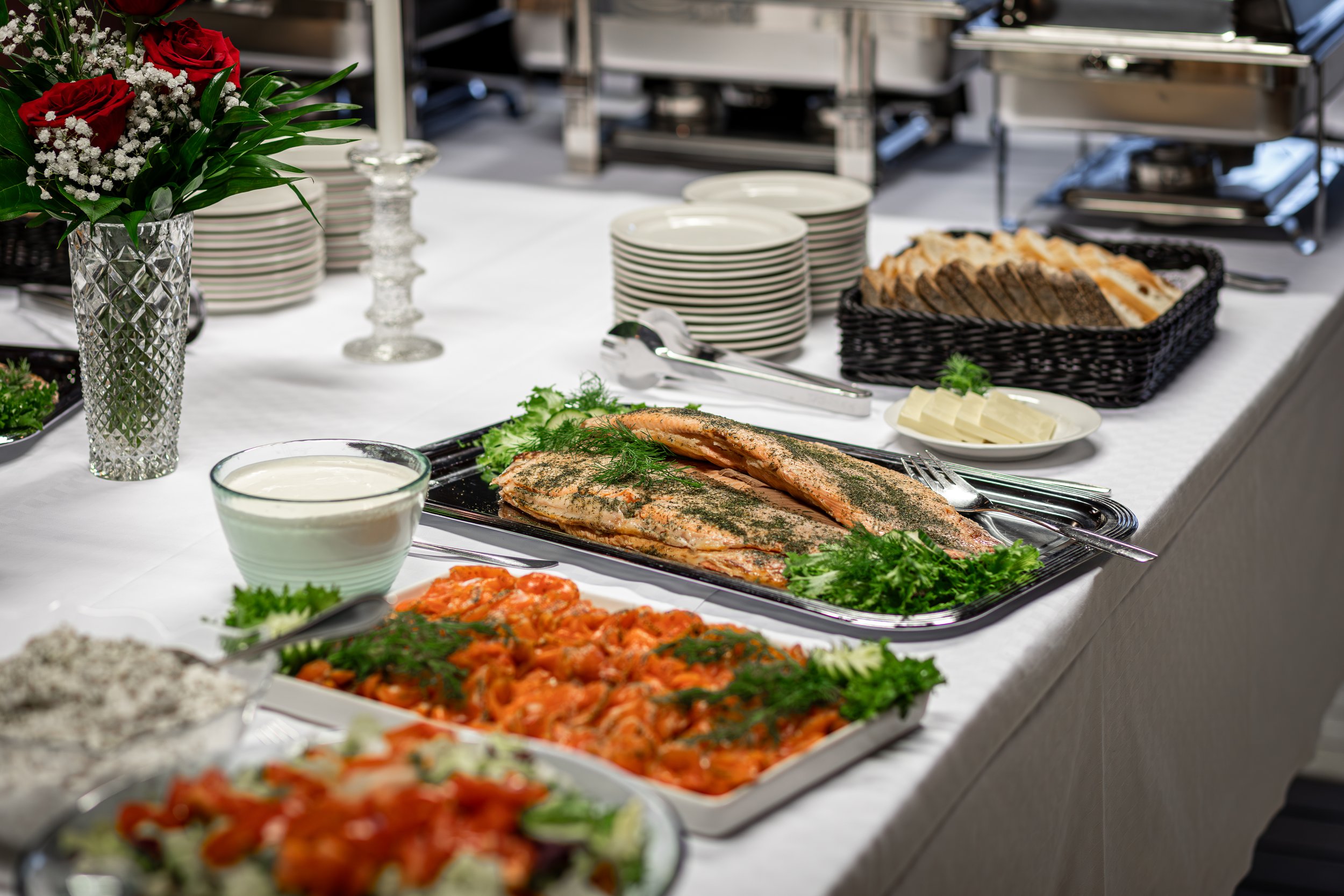 Juhlava pöytä tarjoiltuna ruokaa, sisältäen lohifilee, perunalaatikko, salaattia, juustoa ja leipiä, koristeltuna kukka-asetelmalla.