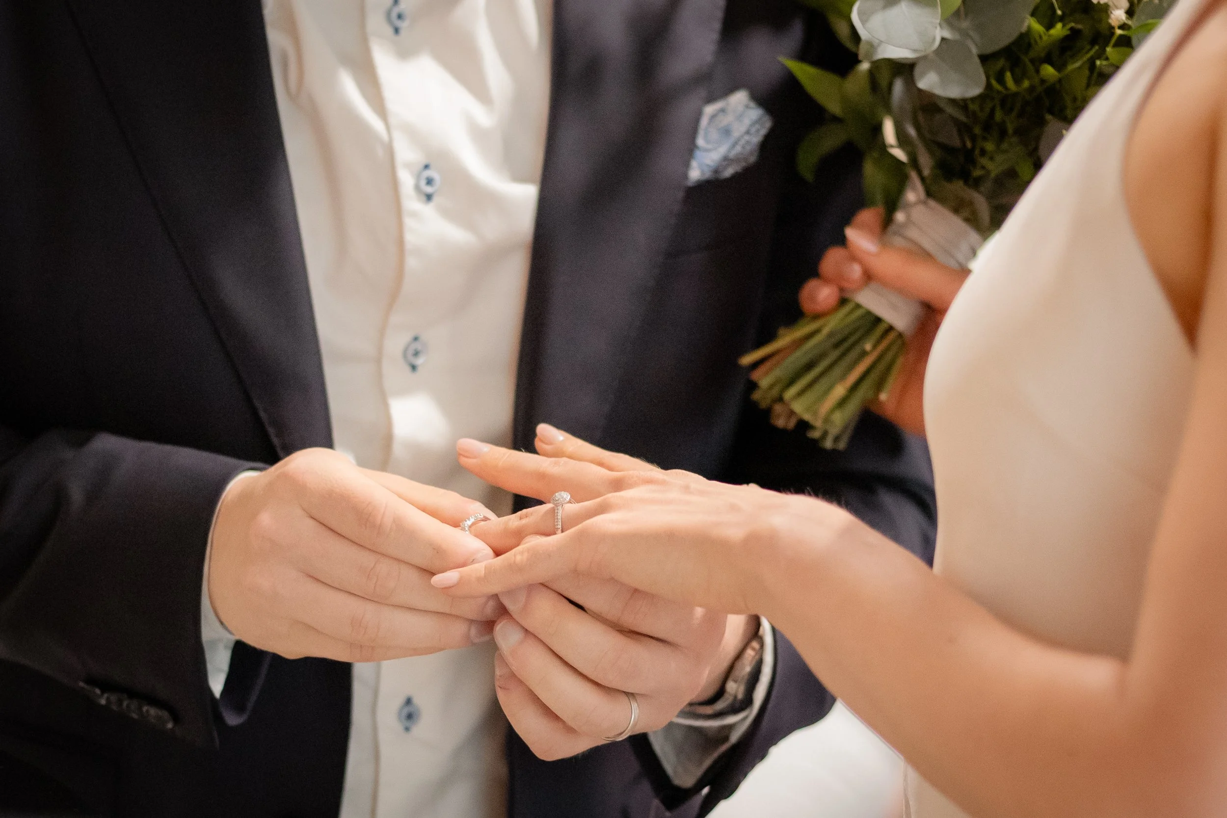 Pariskunta juhlii hääseremonian aikana, mies laittaa sormuksen naisen sormeen ja nainen pitää kukkakimppua käsissään.