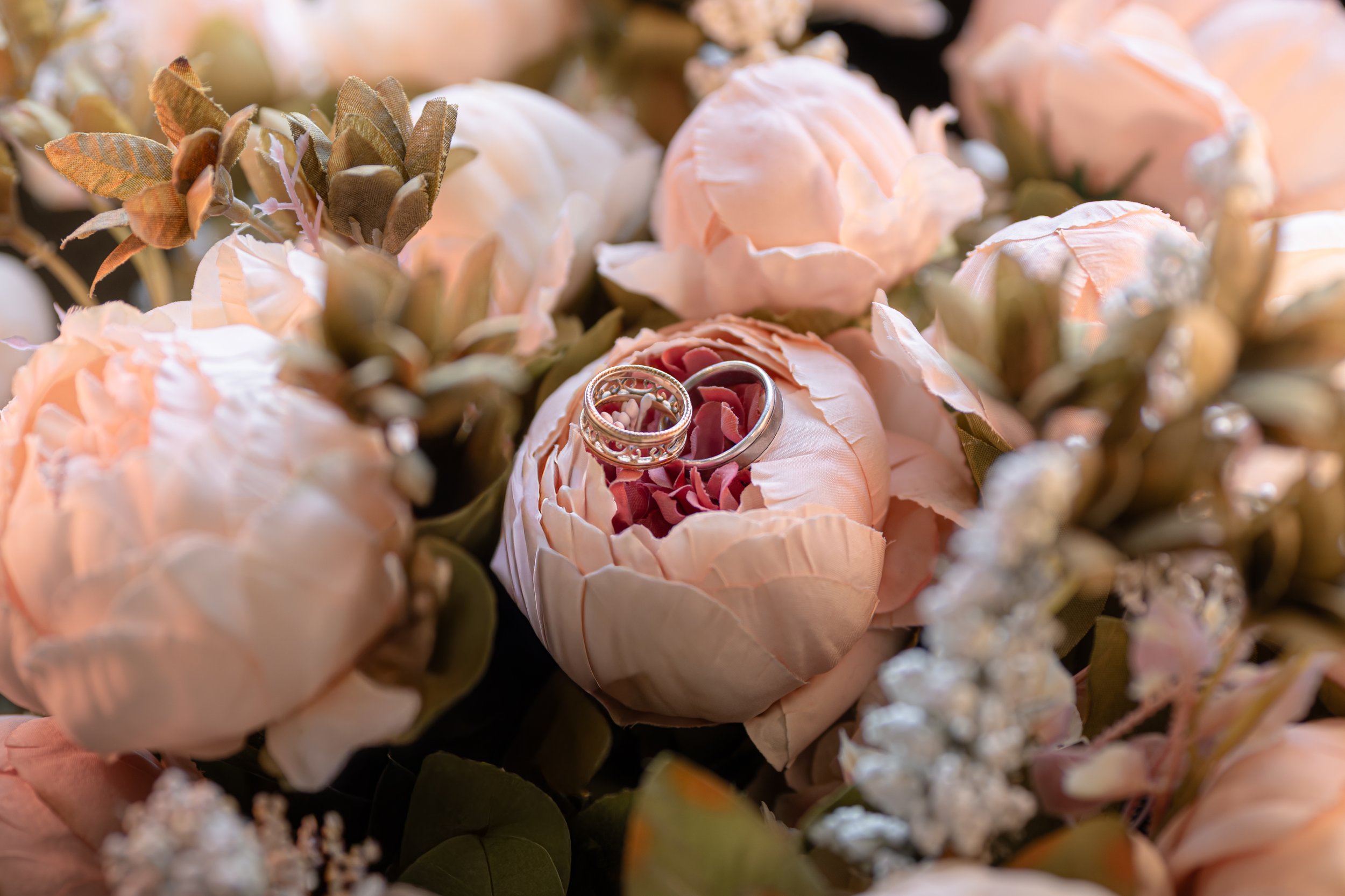 Kuvassa on joukko vaaleanpunaisia kukkia ja vihreitä lehtiä, sekä kaksi kihlasormusta rivissä kukkakedolla.