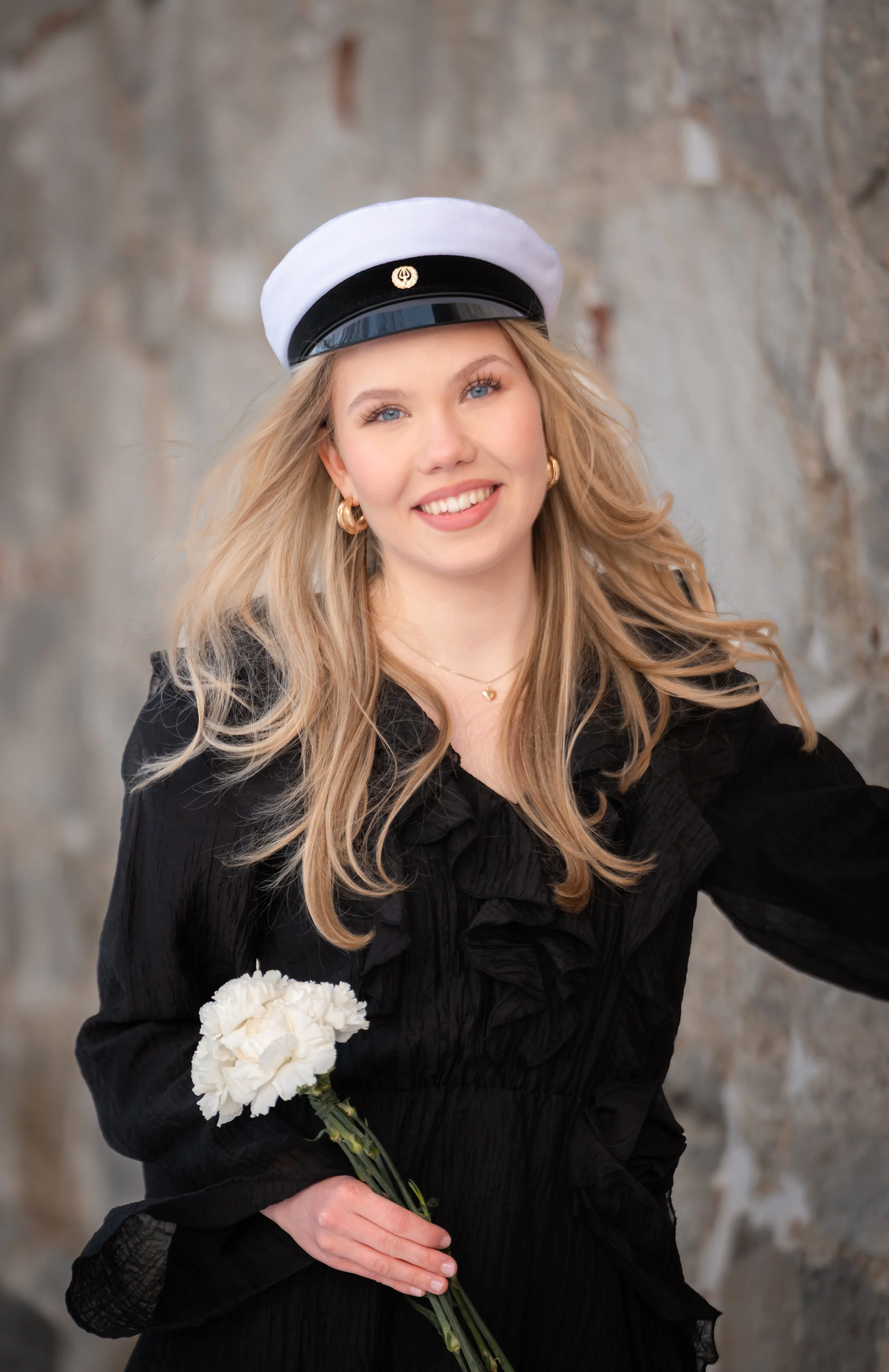 Nuori nainen juhlapuvussa ja valkoisessa ylioppilaslakissa hymyilee ja pitää valkoista narsissikimppua. Taustalla kiviä seinässä.