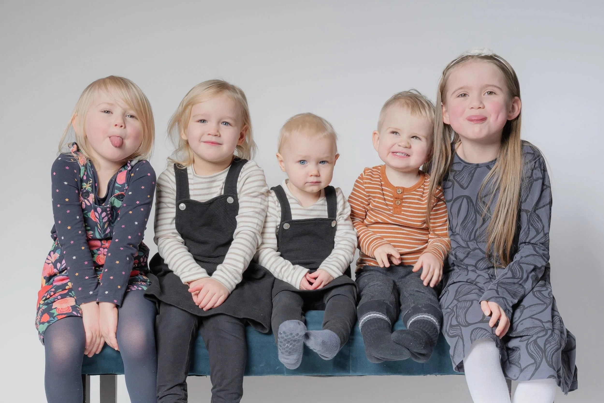 Viisi pikkutyttöä istuu rivissä valkoisella taustalla, hauskoja ilmeitä ja röyhiä kieltoja.