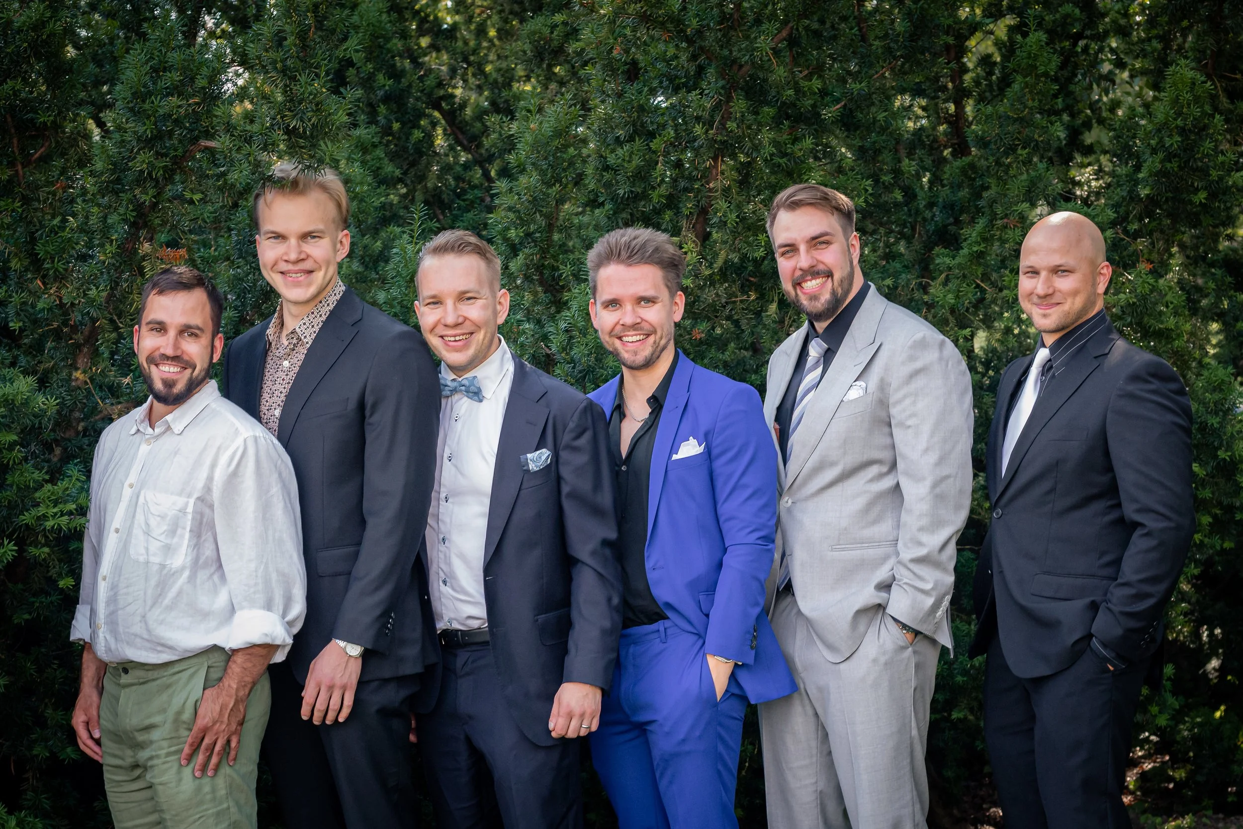 Seitsemän miestä pukuasuissa seisoo viherpensaiden taustalla pihalla hymyillen.