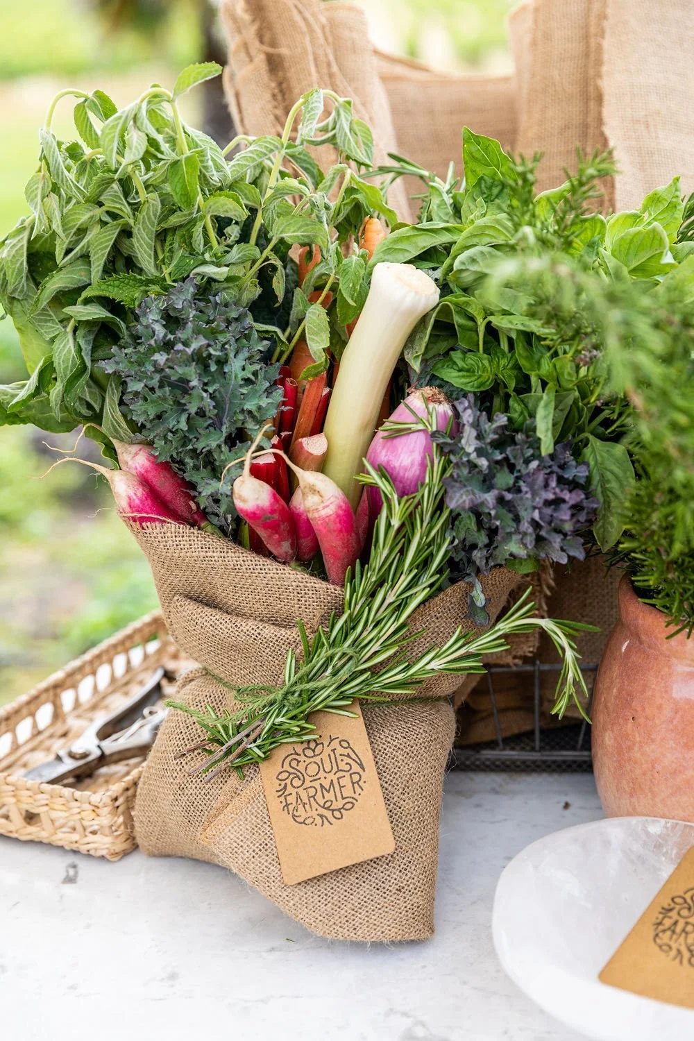 A burlap-wrapped bouquet of fresh garden vegetables and herbs, including radishes, carrots, leeks, onions, rosemary, kale, and basil, with a tag that says 'Soul Farmer'.