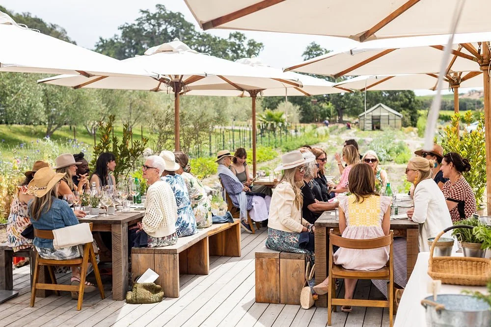 Group of people dining outdoors under large umbrellas on a patio surrounded by greenery.