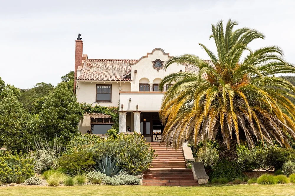 A large house with Spanish-style architecture, surrounded by lush greenery and a tall palm tree, with stairs leading up to the entrance.