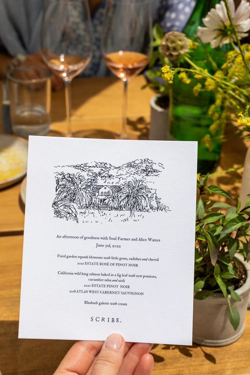 A hand holding a white paper menu with black text and a line drawing of a house surrounded by palm trees, set on a wooden table. The table has glasses, a green vase with flowers, and a plate of food, with a person in a patterned shirt in the backgrou
