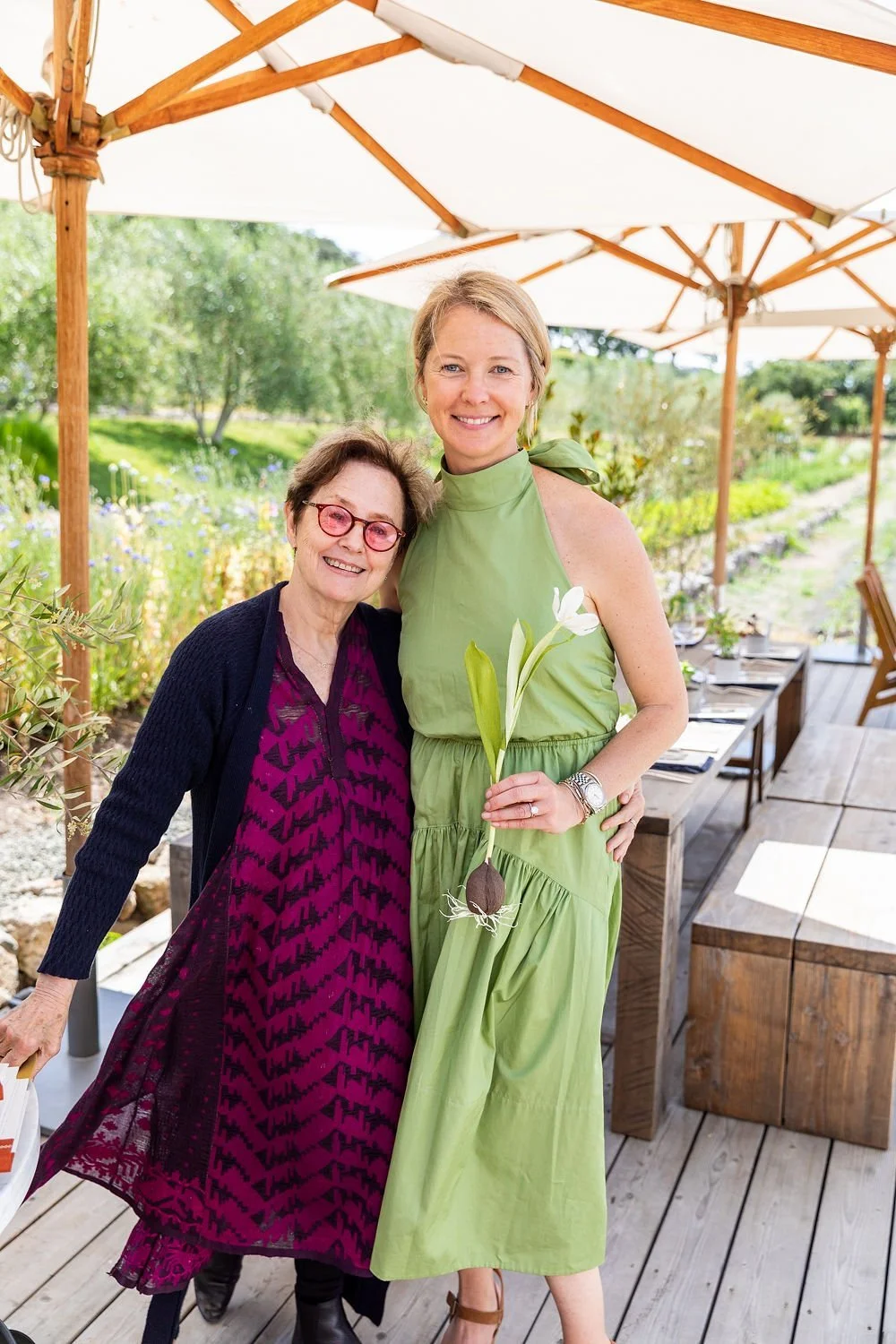 Two women standing outdoors under a large white umbrella, smiling at the camera. The woman on the left is older with short brown hair, red glasses, and wearing a purple patterned dress with a dark cardigan. The woman on the right is younger with blon