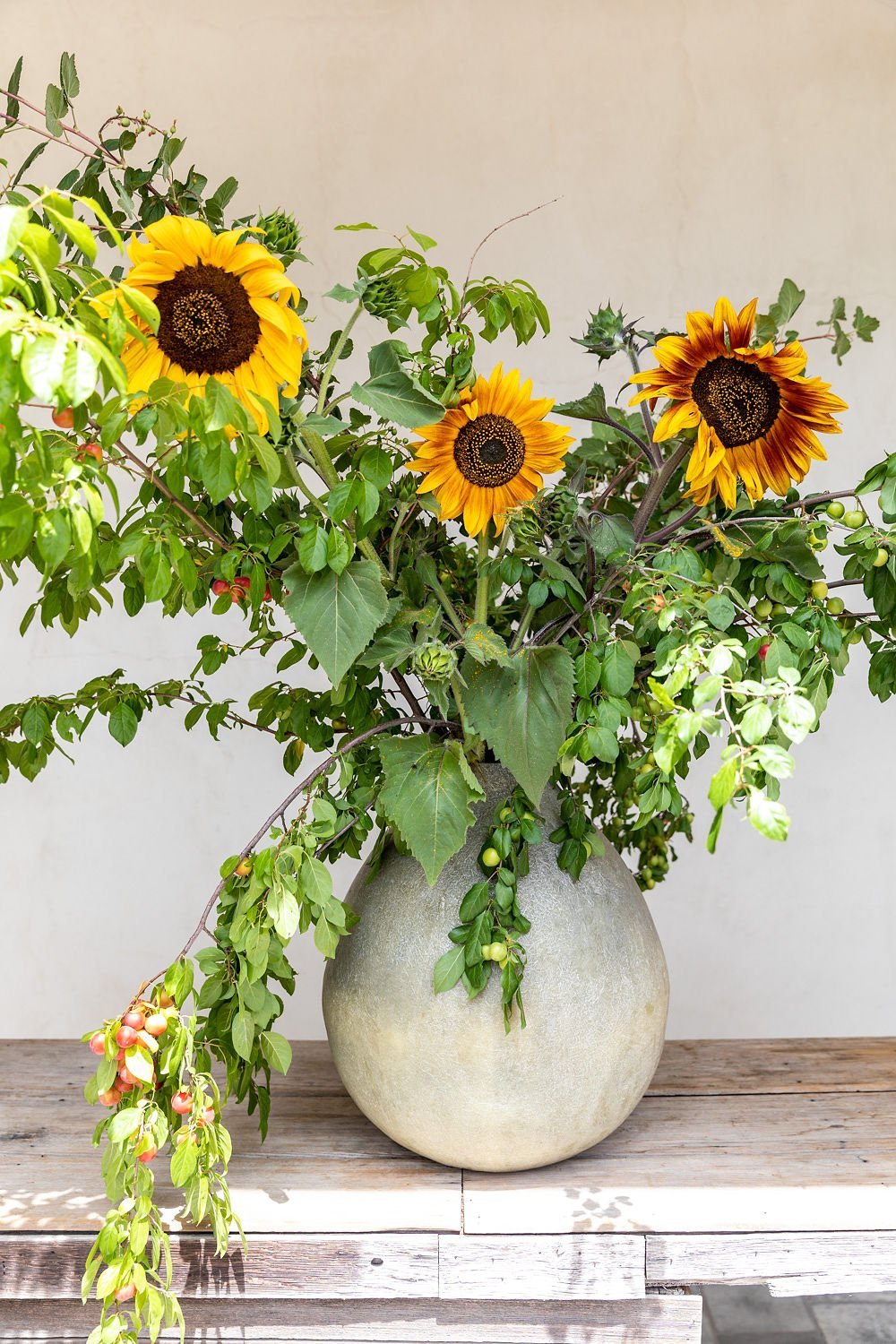 A ceramic vase on a wooden table holds a bouquet of sunflowers, greenery, and blooming branches.