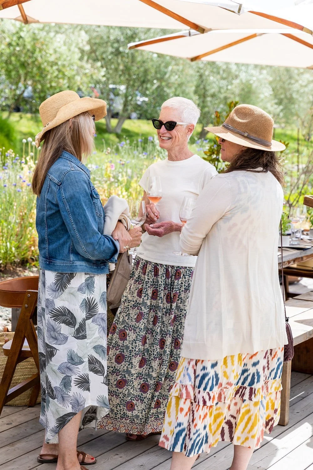 Three women are standing outdoors under large white umbrellas, engaging in conversation and holding glasses of rosé wine. They are dressed in summery outfits with sun hats and sunglasses.