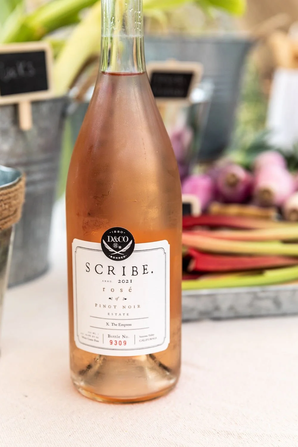 A bottle of Scribe Rosé wine from the 2021 Pinot Noir estate, labeled 'X. The Empress', with a wine bottle number 9309 in Summer Valley, California, placed on a table with blurred vegetables and produce in the background.