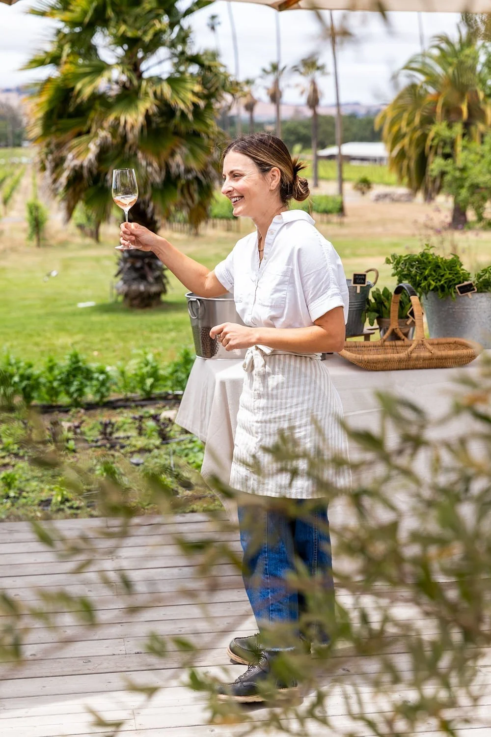 A woman in a white shirt and apron holding a glass of rosé wine outdoors, smiling in a garden with palm trees and greenery.