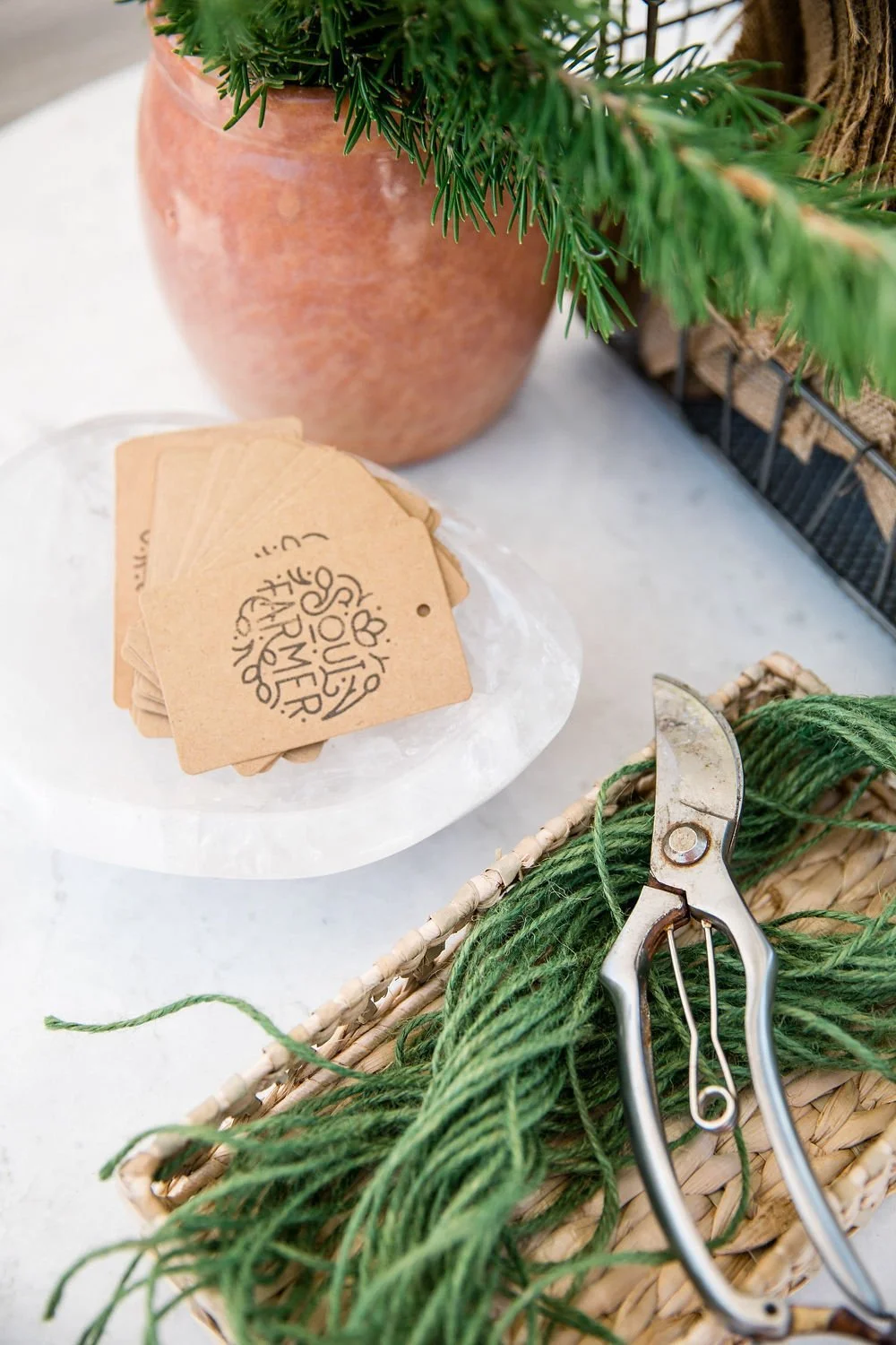 A pair of pruning shears, green pine branches, a basket, a potted plant with green foliage, and kraft paper tags with handwritten text on a white surface.